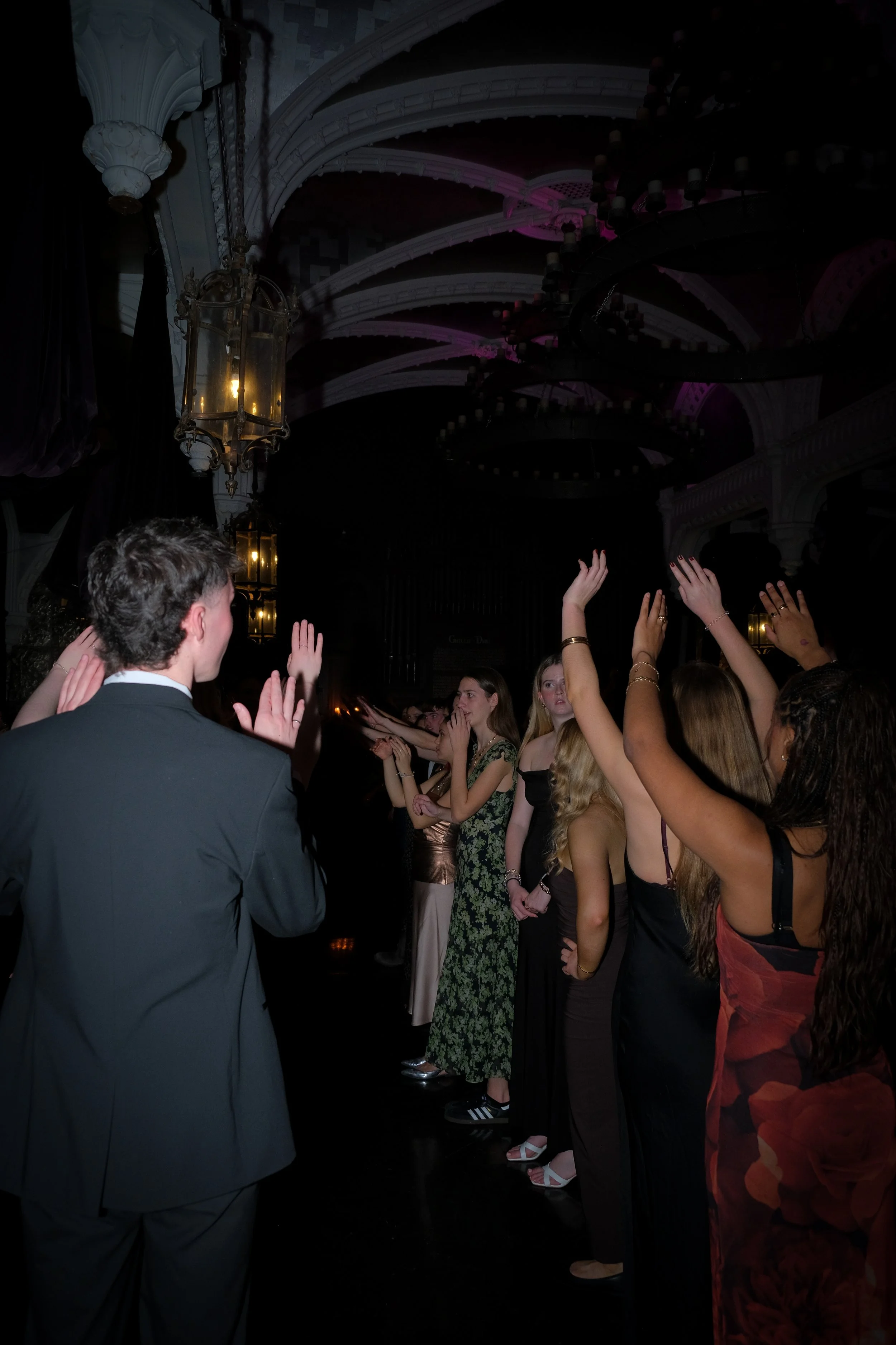 People dancing and raising their hands in a dark, elegant venue with ornate ceiling and chandeliers.