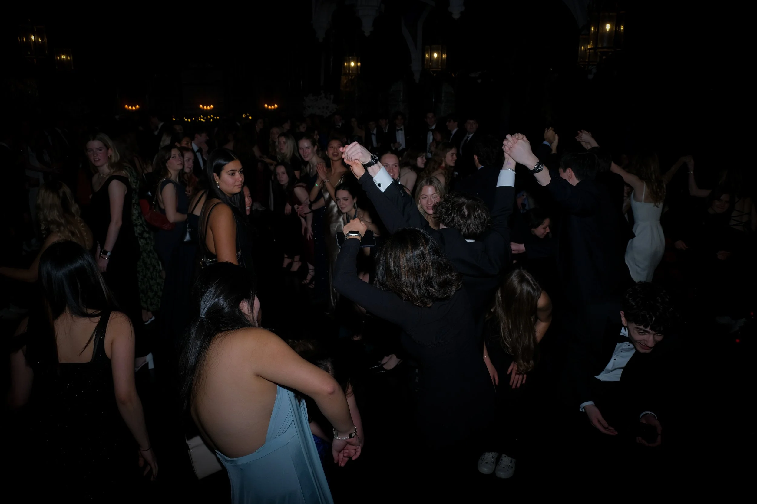 A group of young people dancing at a formal event or party, many dressed in elegant attire. Some are dancing closely, while others observe or dance in the background, in a dimly lit setting with warm lighting.