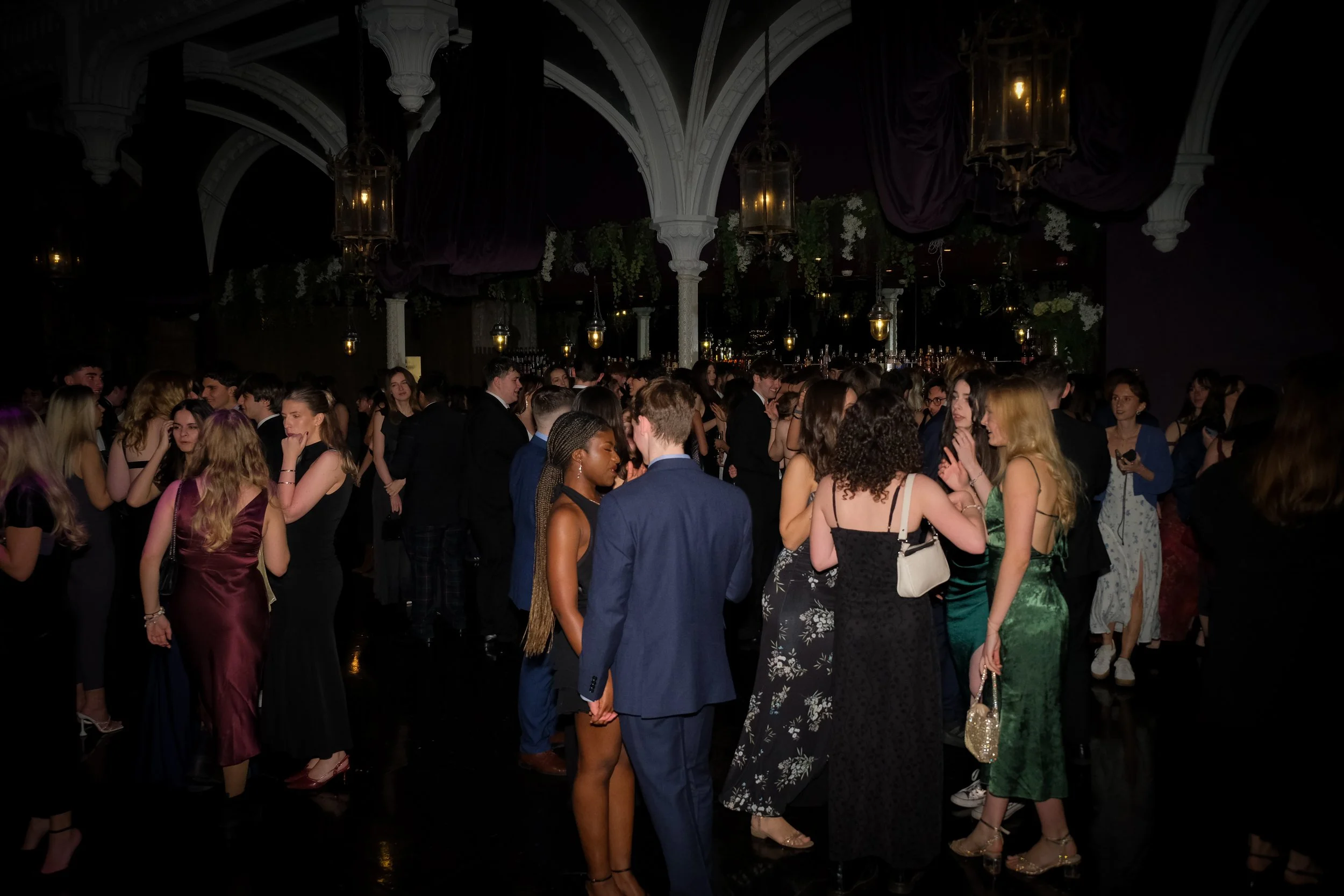 People dancing and socializing at a formal indoor event or party, with ornate architecture and dim lighting.
