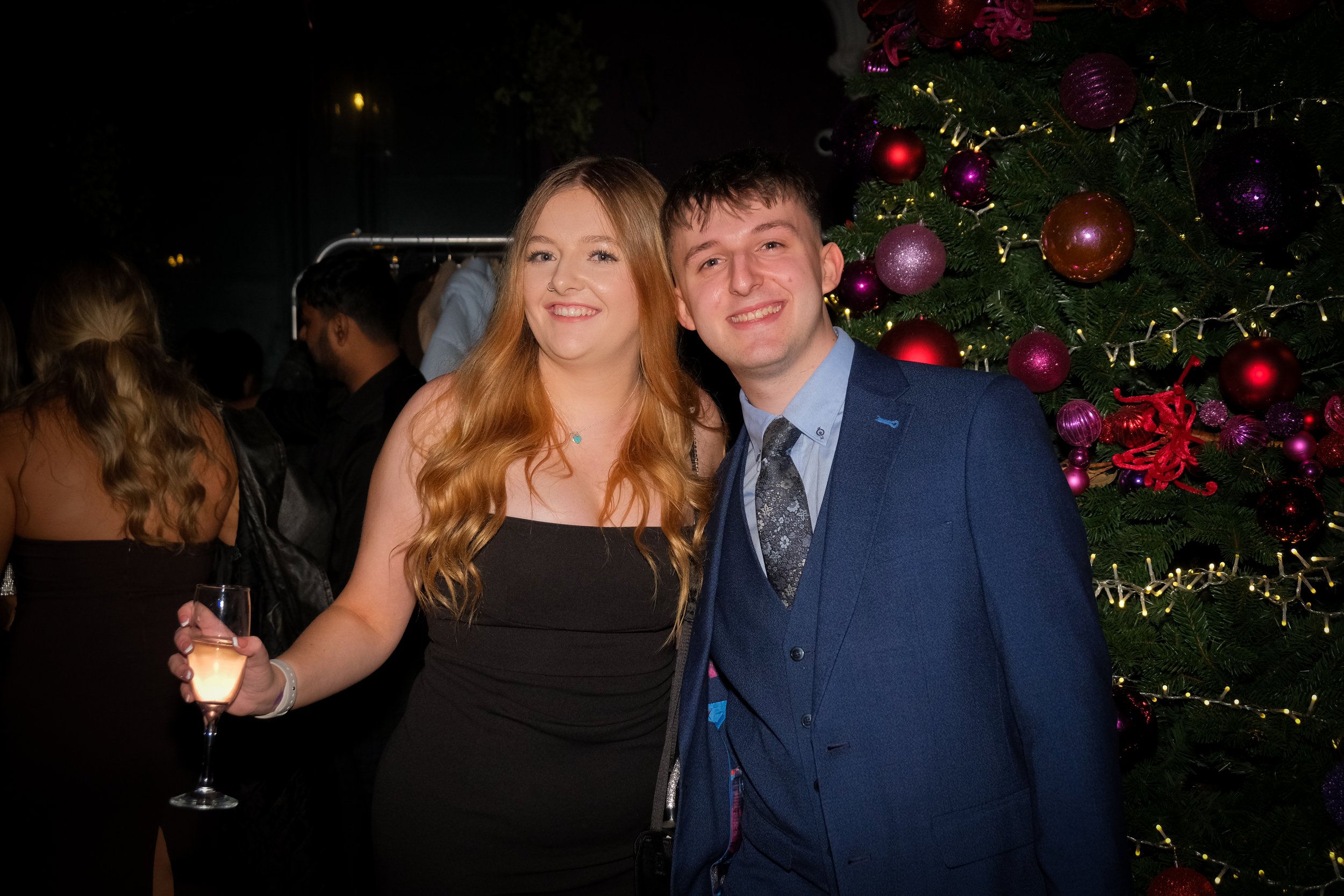 A young woman with long red hair wearing a black dress and holding a glass of champagne, standing next to a young man in a blue suit and tie, both smiling, in front of a decorated Christmas tree with purple and red ornaments and lights at a holiday c