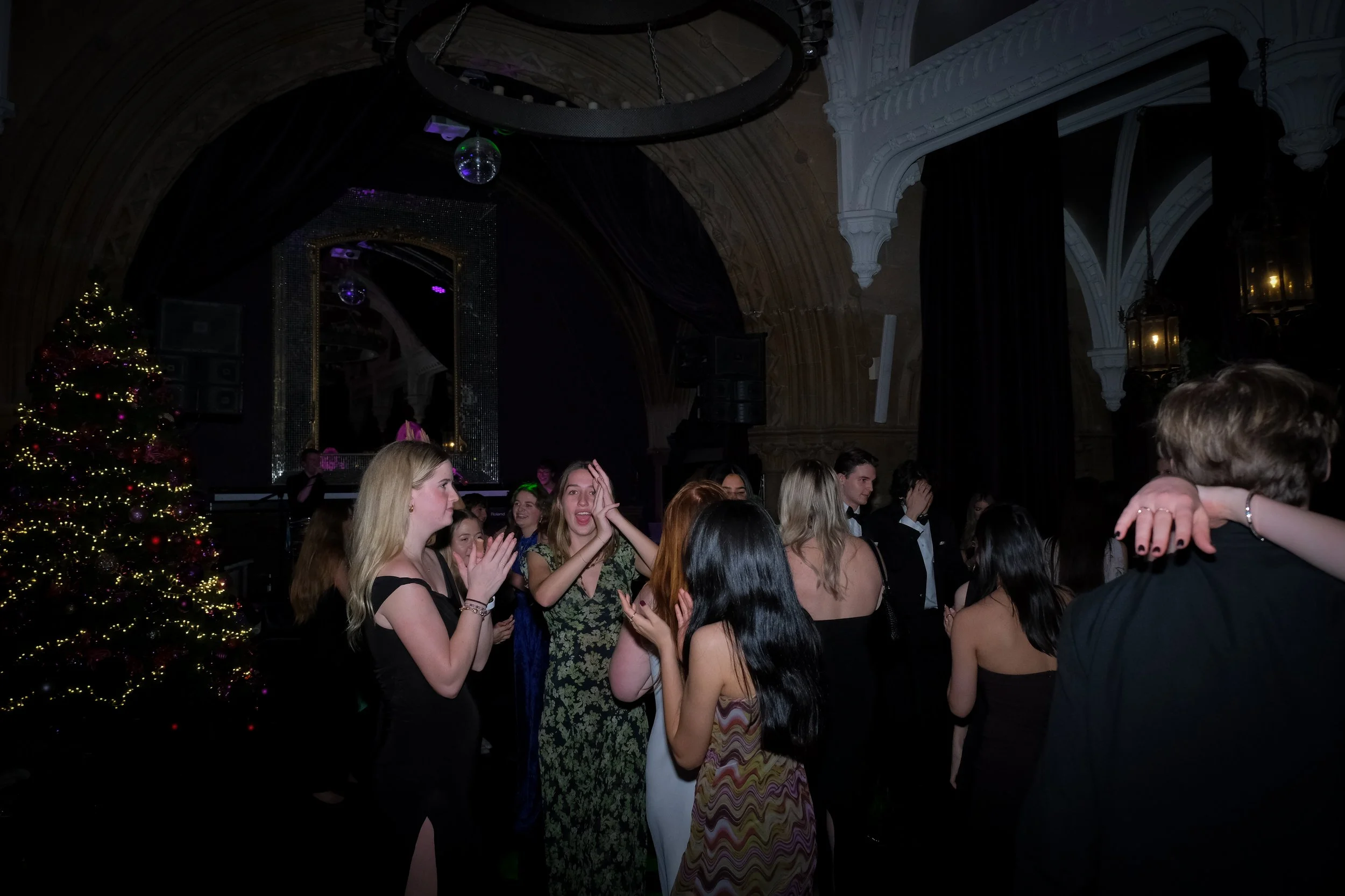 People dancing and socializing at a party with a decorated Christmas tree, a large mirror, and gothic-style architecture in the background.