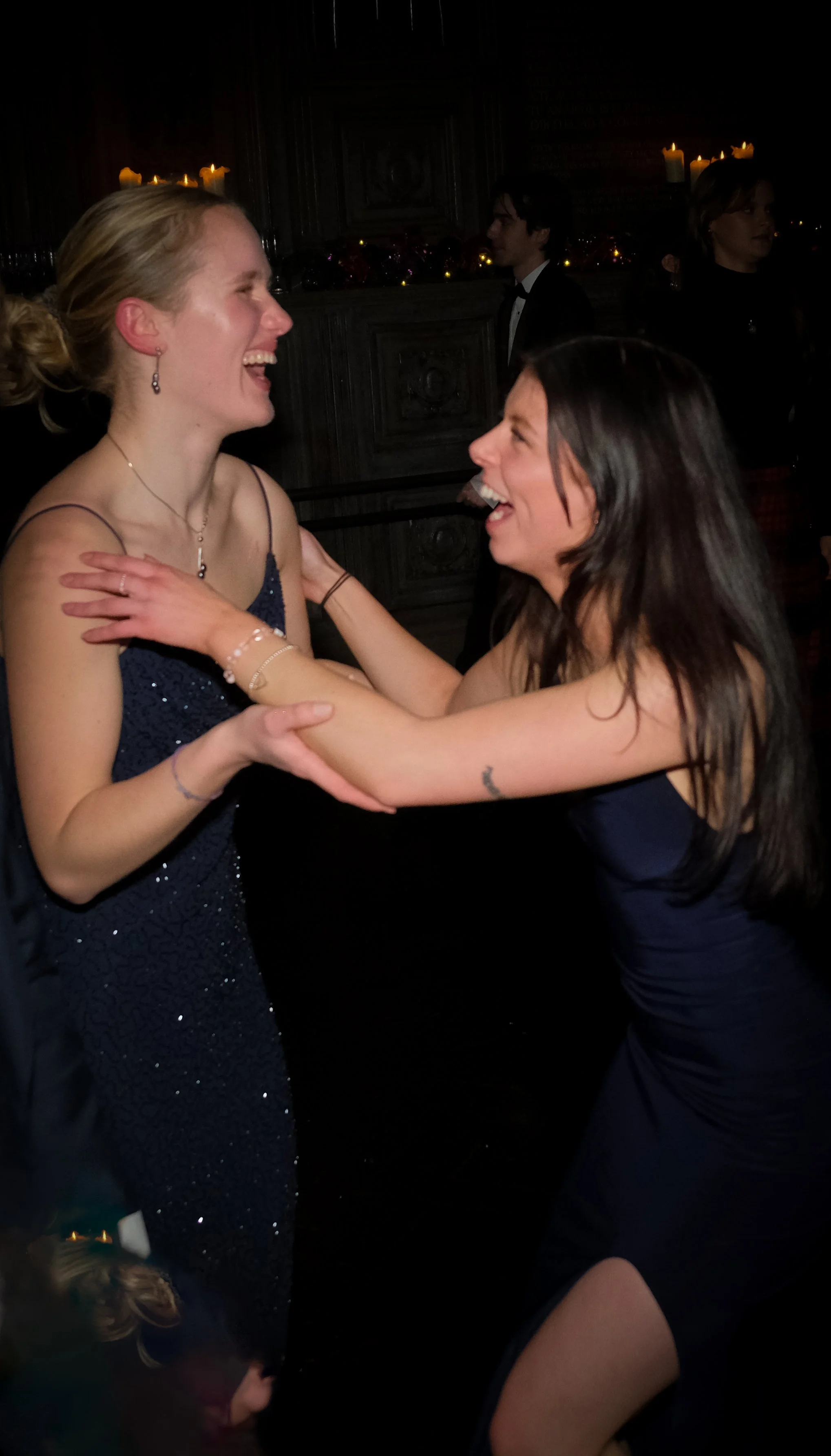 Two women in elegant dresses laughing and embracing at a celebration or party with candles in the background.
