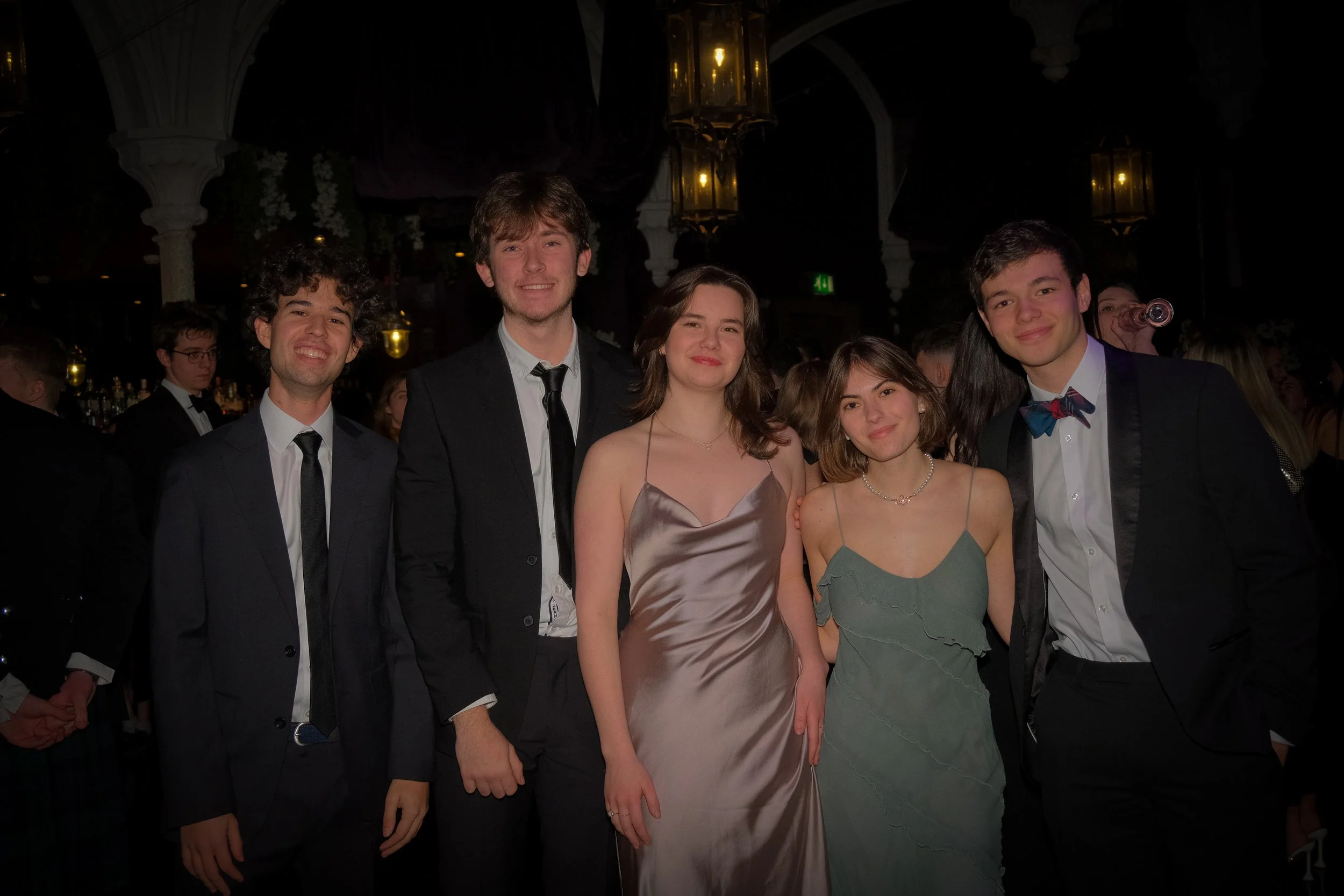 Group of five young people in formal attire at a social event or dance, standing together and smiling, with a dark background and elegant lighting.
