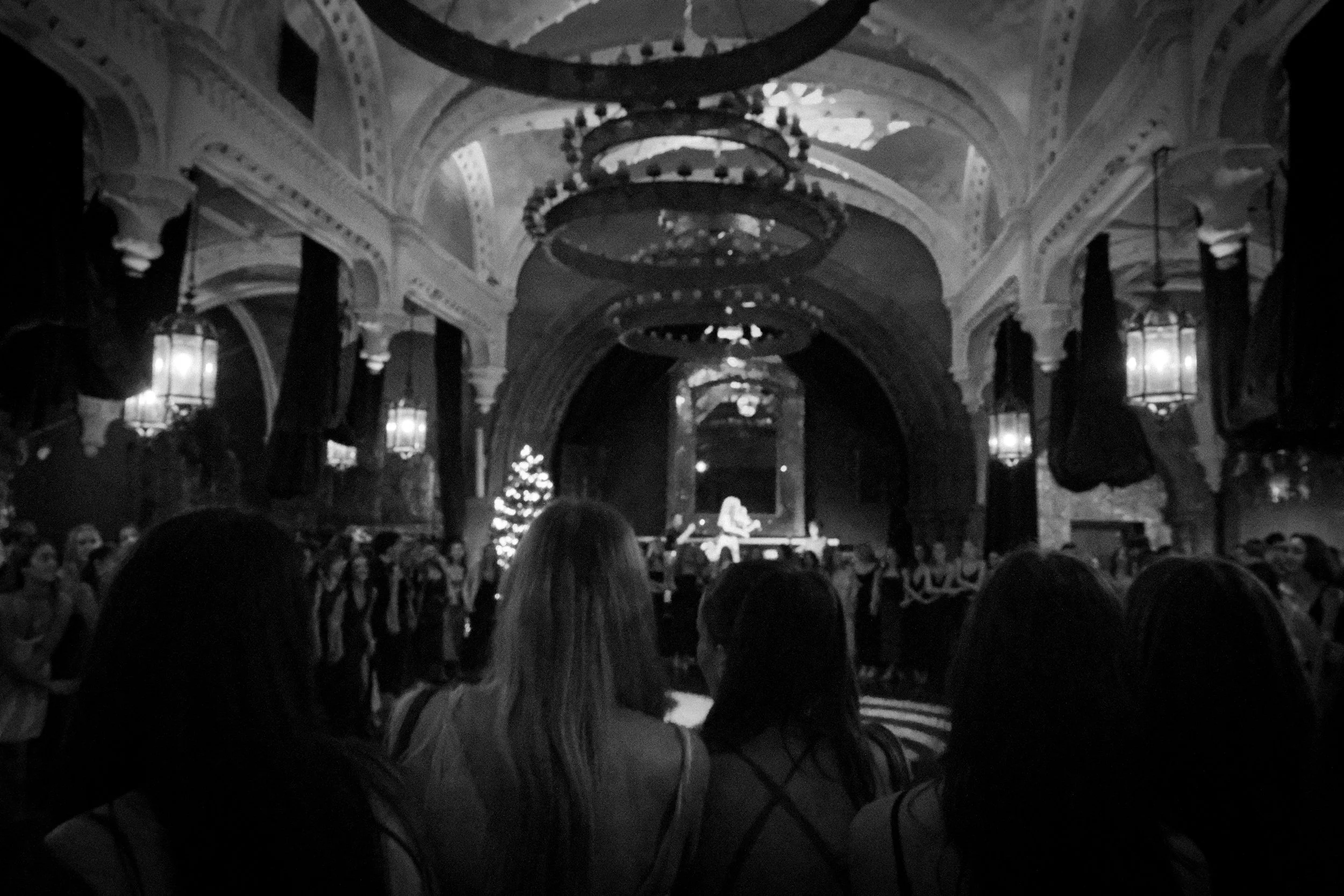 A live musical performance in an ornate hall with an arched ceiling decorated with chandeliers, people watching the performer, and a Christmas tree on the side.