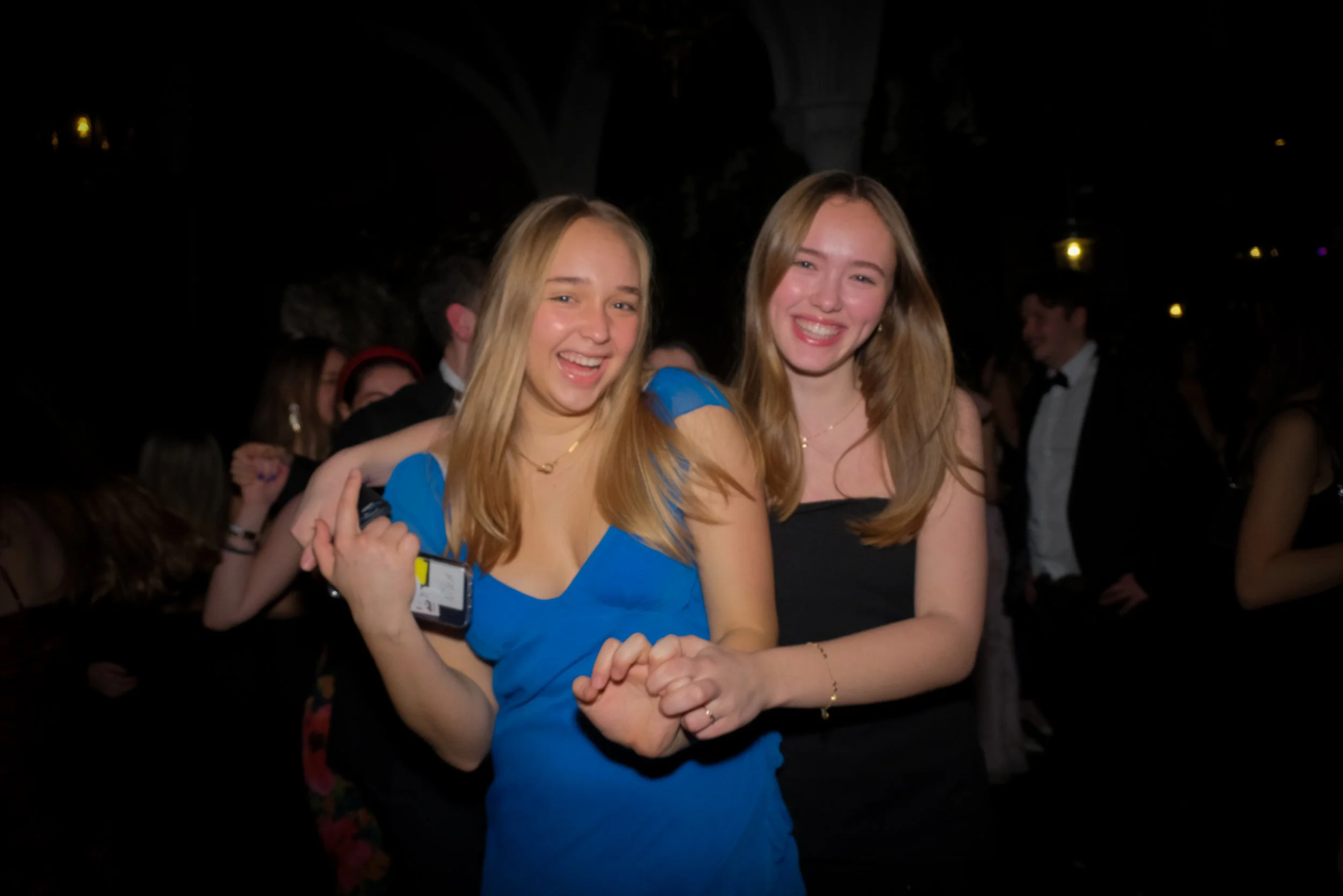 Two young women with long hair smiling and dancing together at a party, one wearing a blue dress and the other a black dress, with other people in formal attire in the background.
