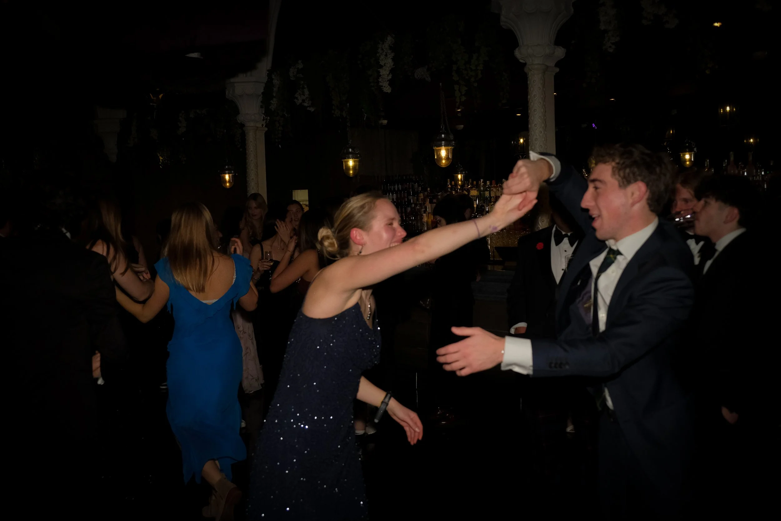 A young woman and young man dancing and holding hands at a formal event, surrounded by other guests in elegant attire, in a dimly lit venue with hanging lights and decorative columns.