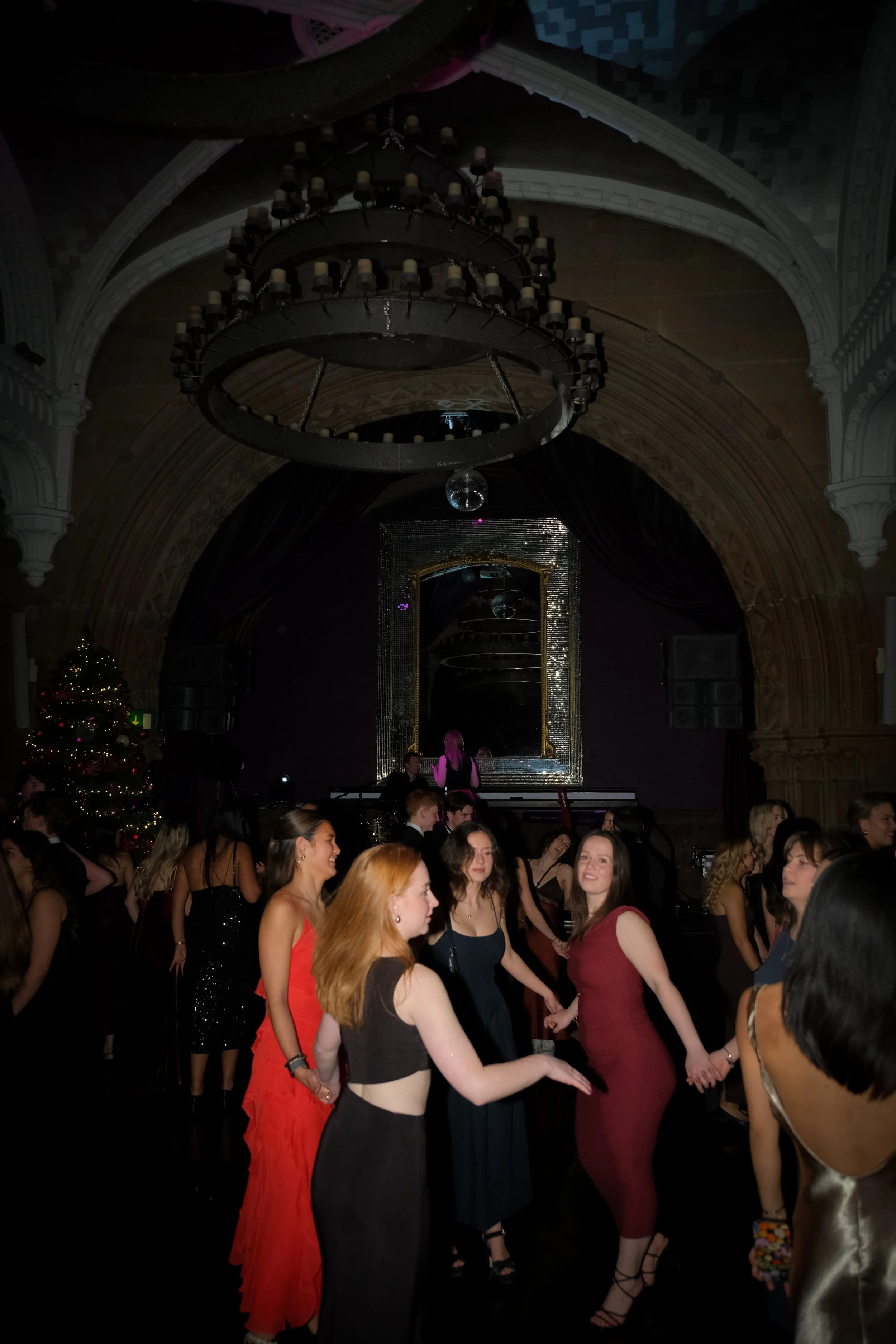 People dancing at a party with a stage, large chandelier, and a Christmas tree in the background.