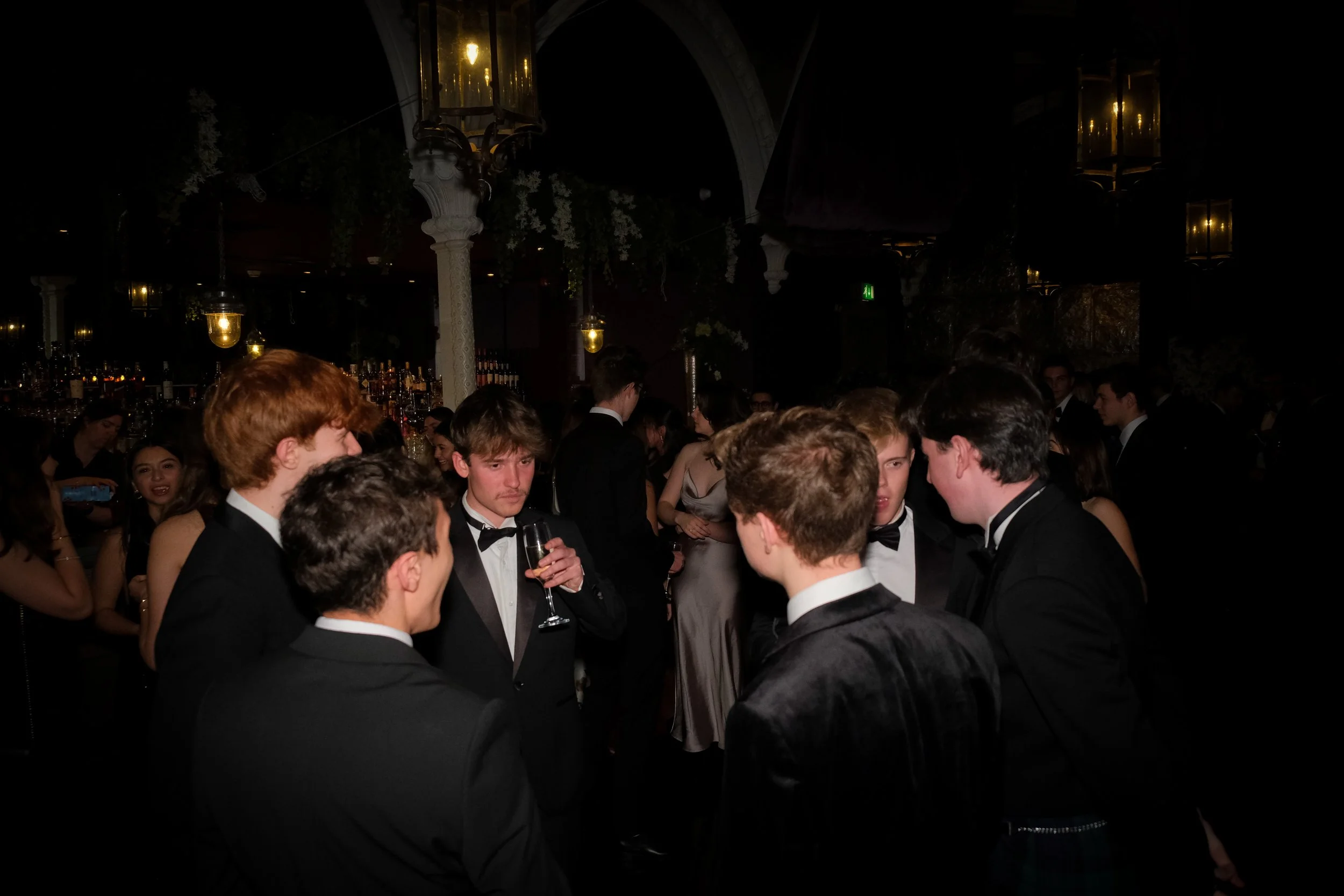 Group of young men in tuxedos socializing at a formal event or black-tie party in a dimly lit venue with hanging lights and a bar in the background.