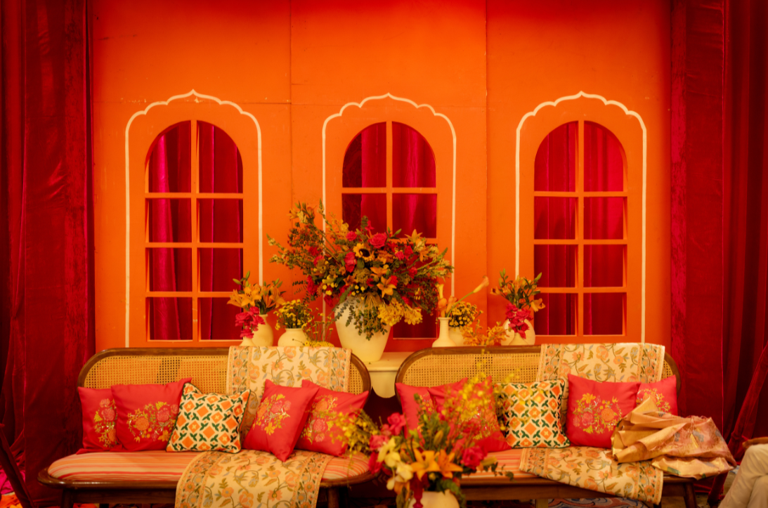 A decorative indoor setting with coral-colored walls, three large window-like shapes with white borders, and red curtains. There is a floral arrangement with yellow, red, and green flowers on a white table, and a sofa with colorful pillows in red, or