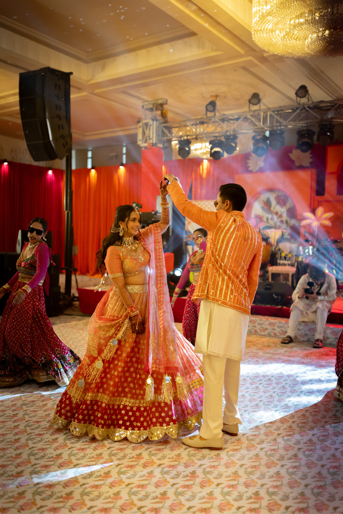 Couple dancing at an Indian wedding reception with colorful traditional attire, surrounded by other guests, stage lighting, and festive decorations.