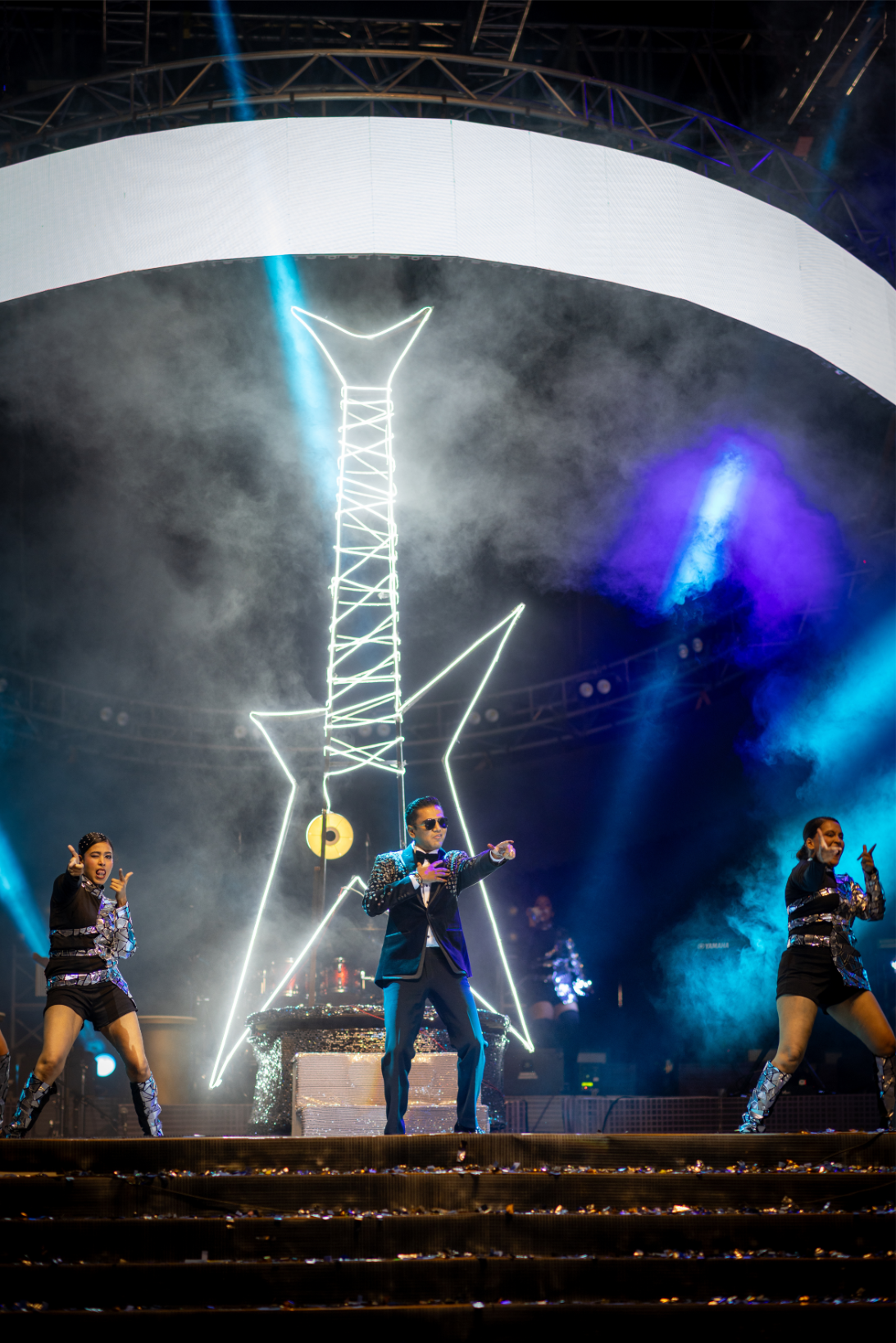 Performer in a black suit and sunglasses dancing on stage with backup dancers, with a bright star-shaped neon light and large screen behind him.