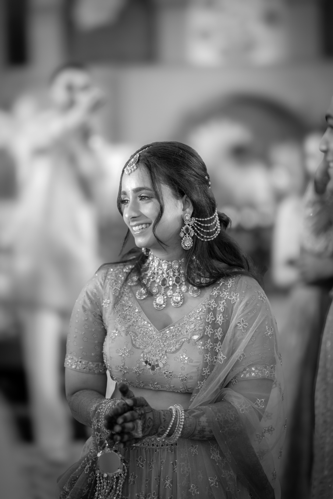 A smiling woman dressed in traditional Indian wedding attire, wearing elaborate jewelry and henna on her hands, at a wedding celebration.