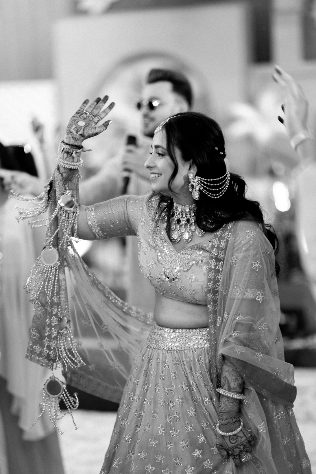 A woman dressed in traditional Indian attire, smiling and waving, with jewelry and henna on her hand, at a celebration or wedding.