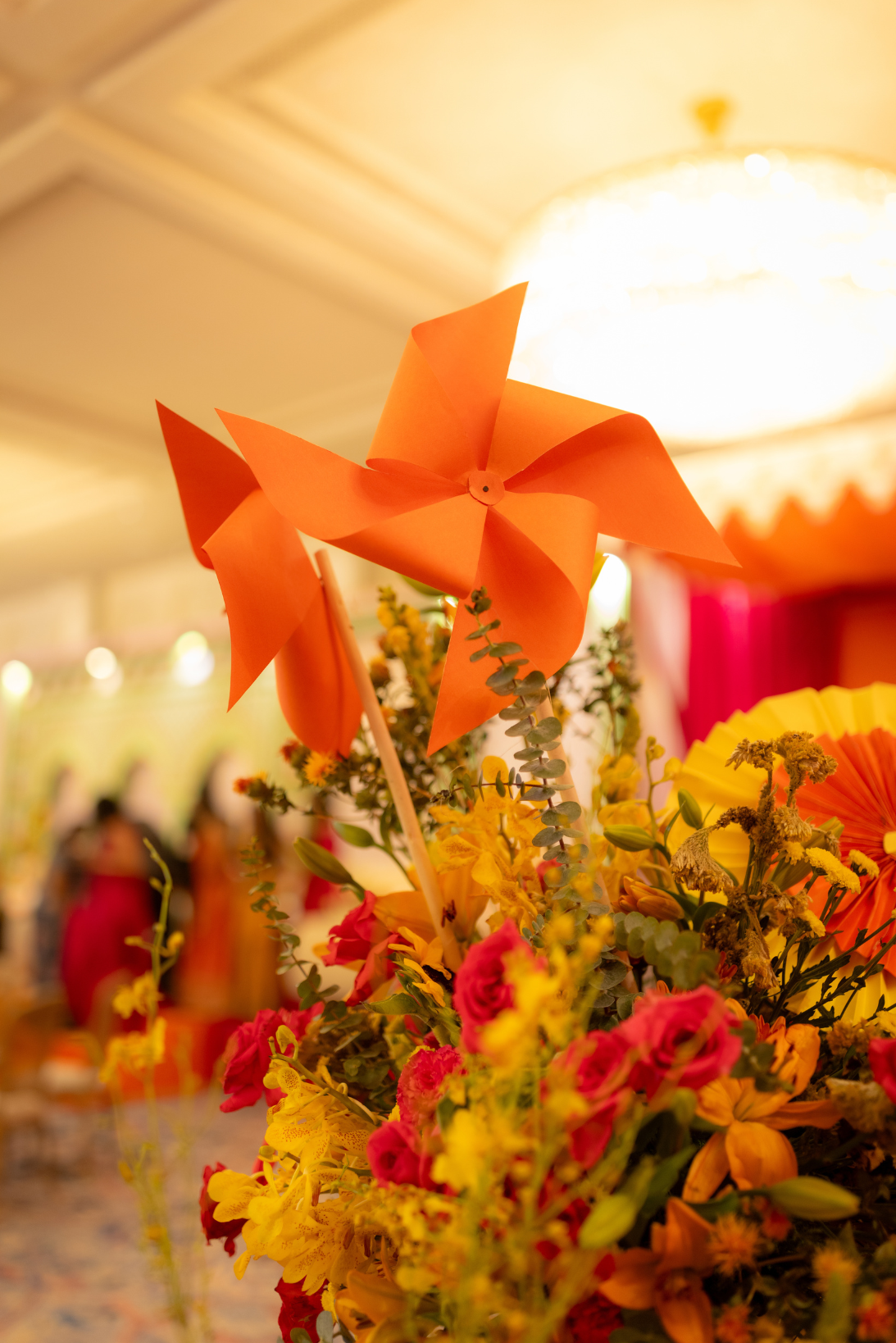 Orange paper pinwheel on floral arrangement with yellow, pink, and orange flowers at a festive event.
