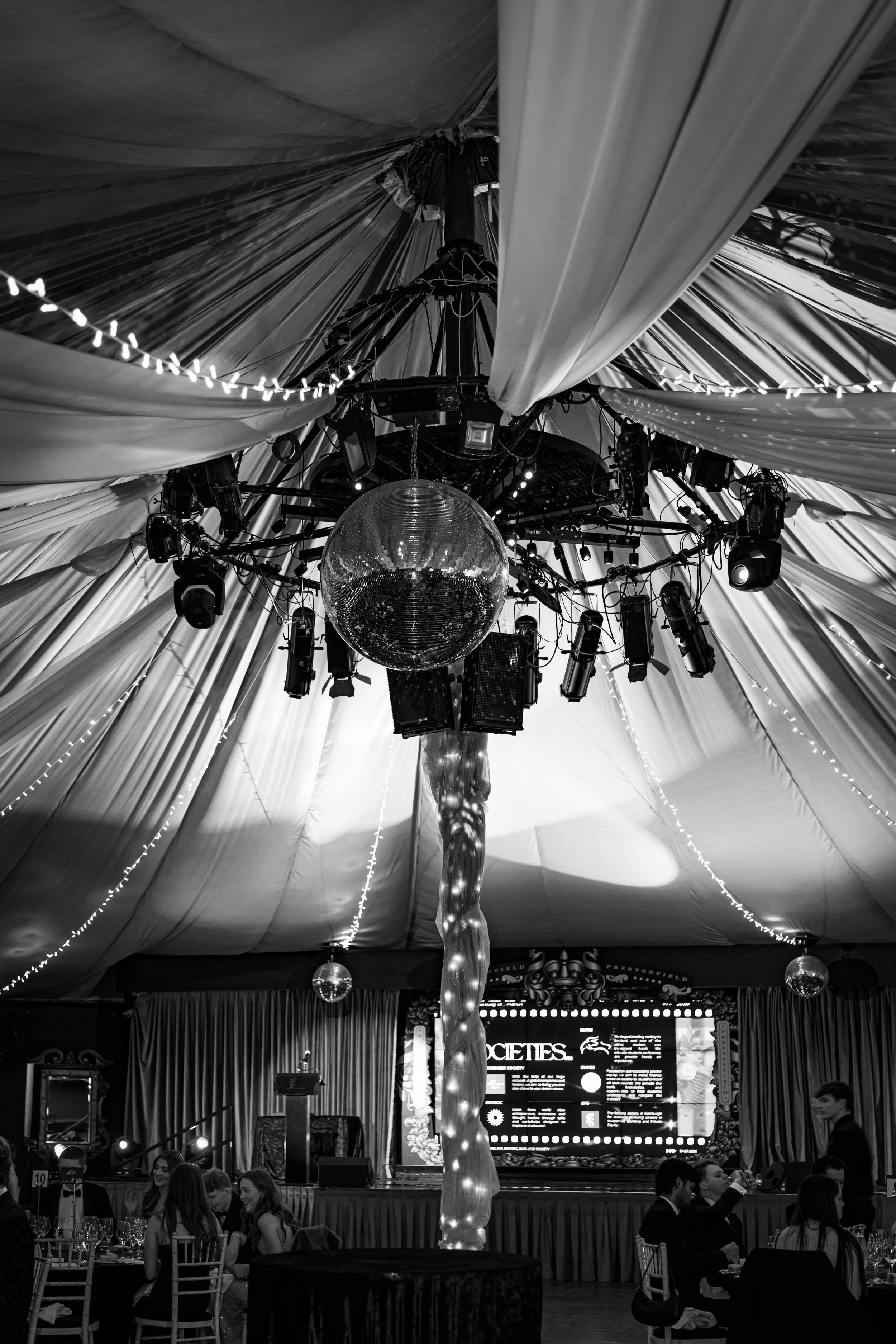 A black and white photo of a large decorated event hall with a wide ceiling draped in fabric. In the center, a disco ball hangs from the ceiling, surrounded by stage lights and sound equipment. People are seated at tables with white chairs, and a lar