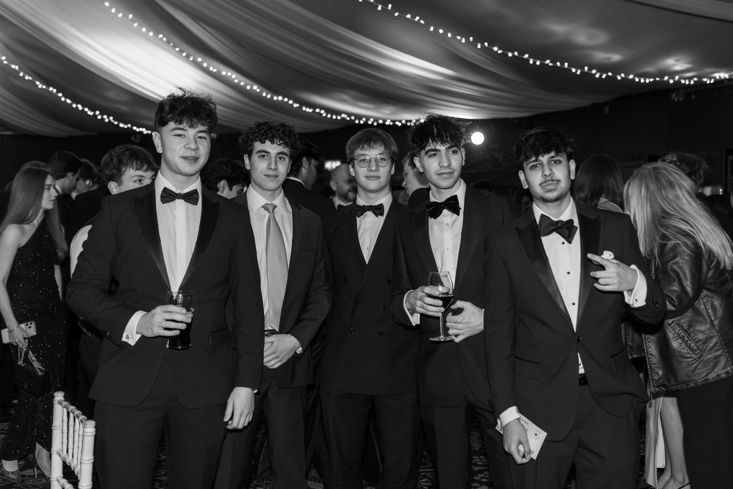 A group of five young men in tuxedos with bow ties standing together at a formal event, holding drinks, with other guests in the background.