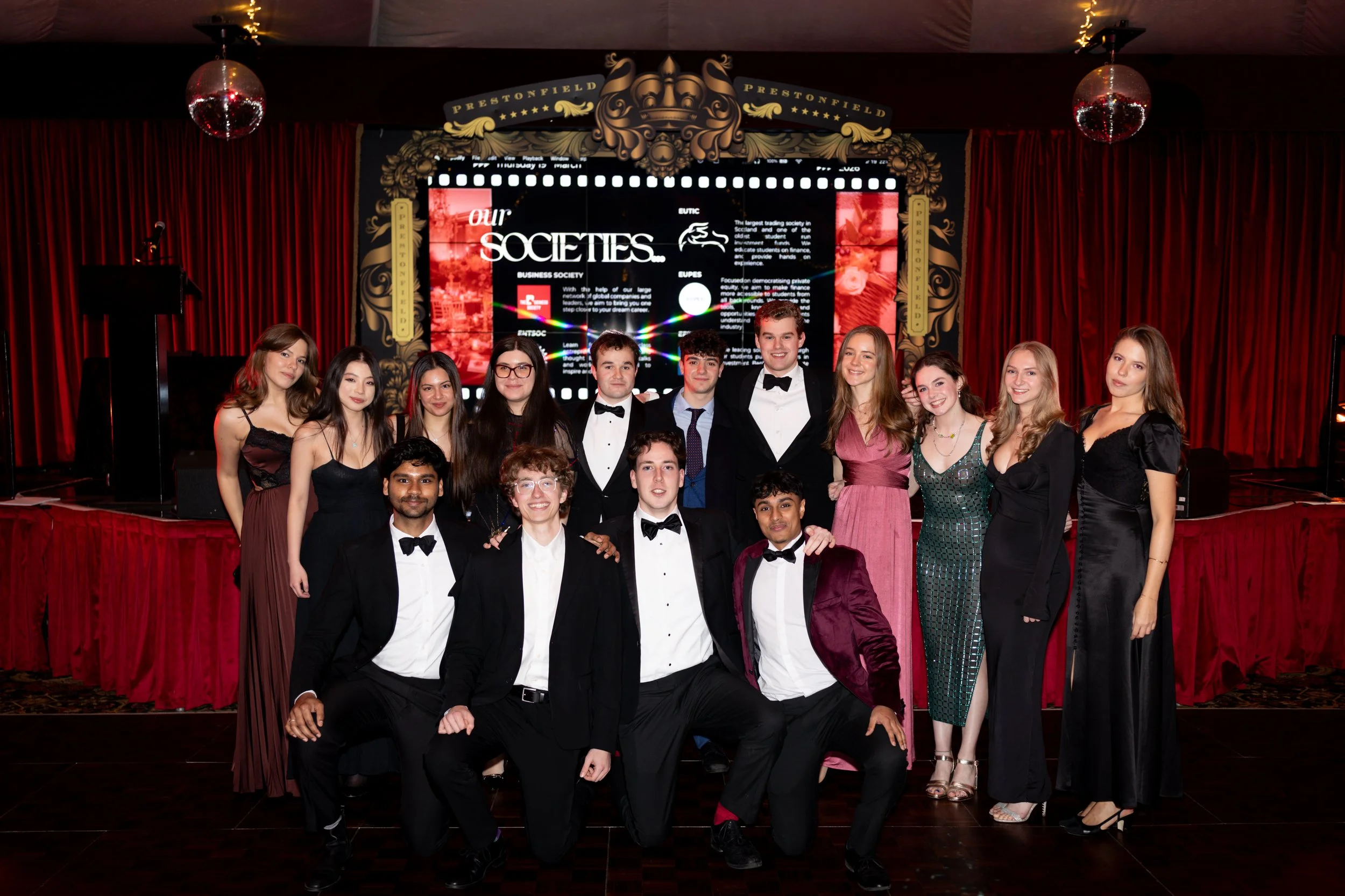 A group of young people dressed in formal attire posing on a stage with red curtains and decorative framing, with a large digital screen displaying text and graphics behind them.