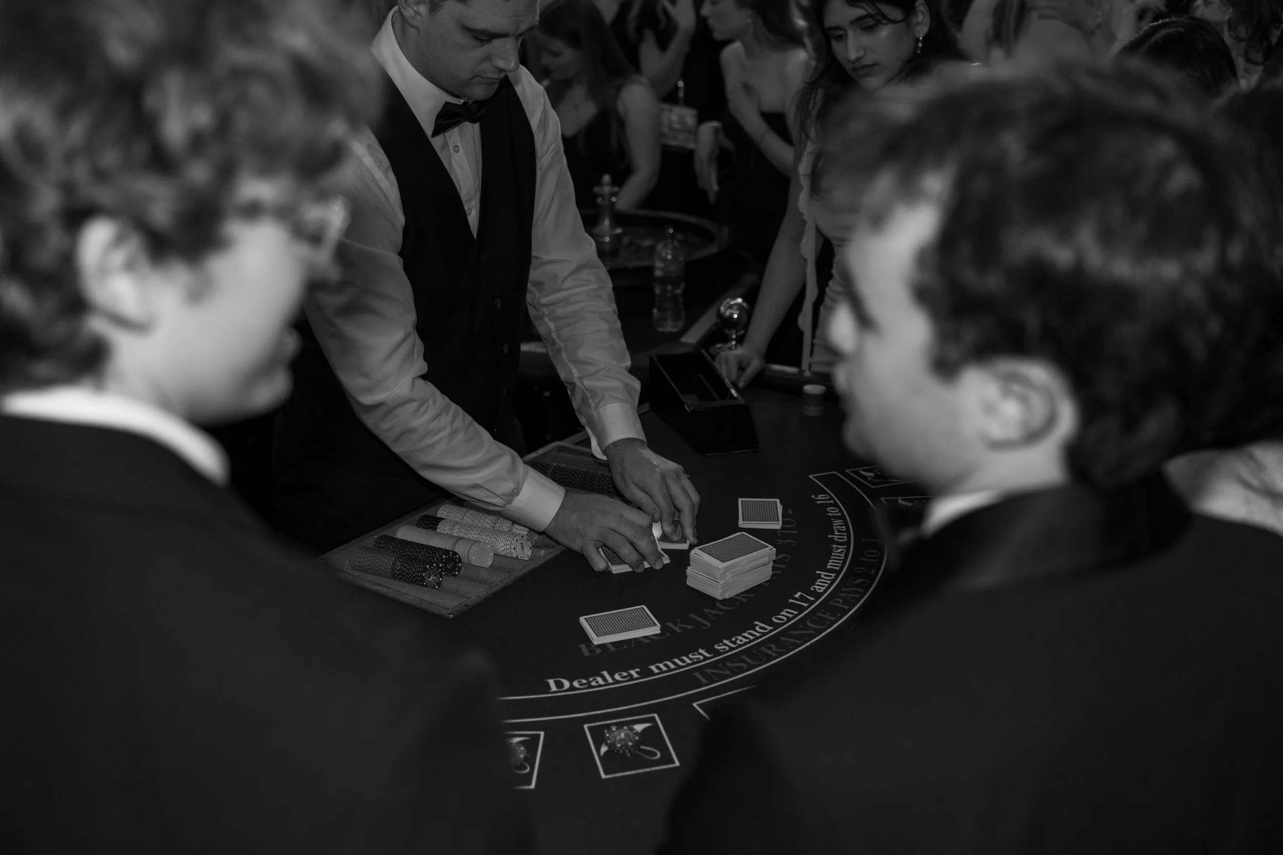 Black-and-white photo of a casino dealer in a tuxedo with a bow tie, dealing cards to young male players dressed in suits, surrounded by other guests at the table.