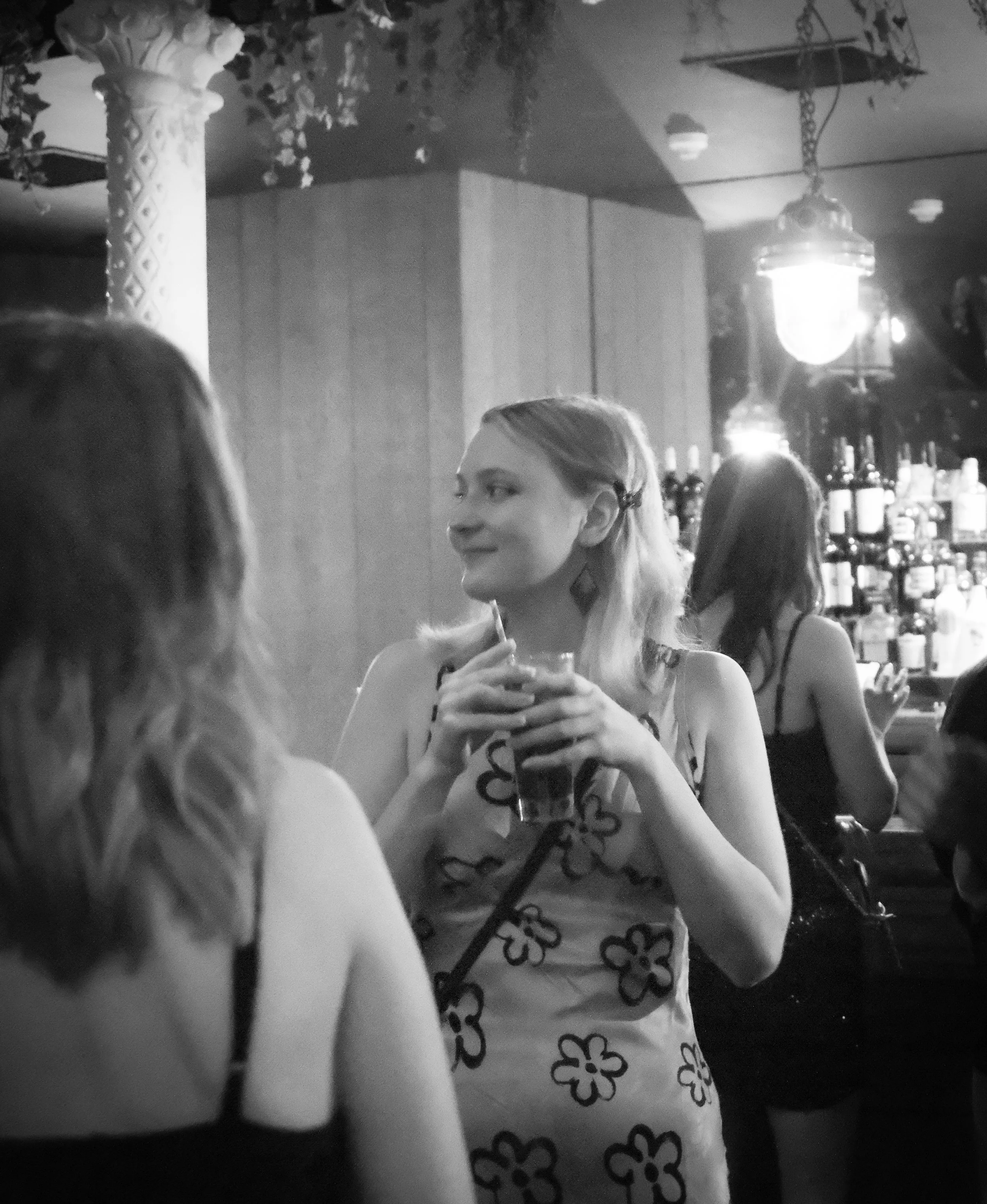 A woman in a floral dress holding a drink inside a bar, with other women in the background, and bottles of alcohol behind the bar.