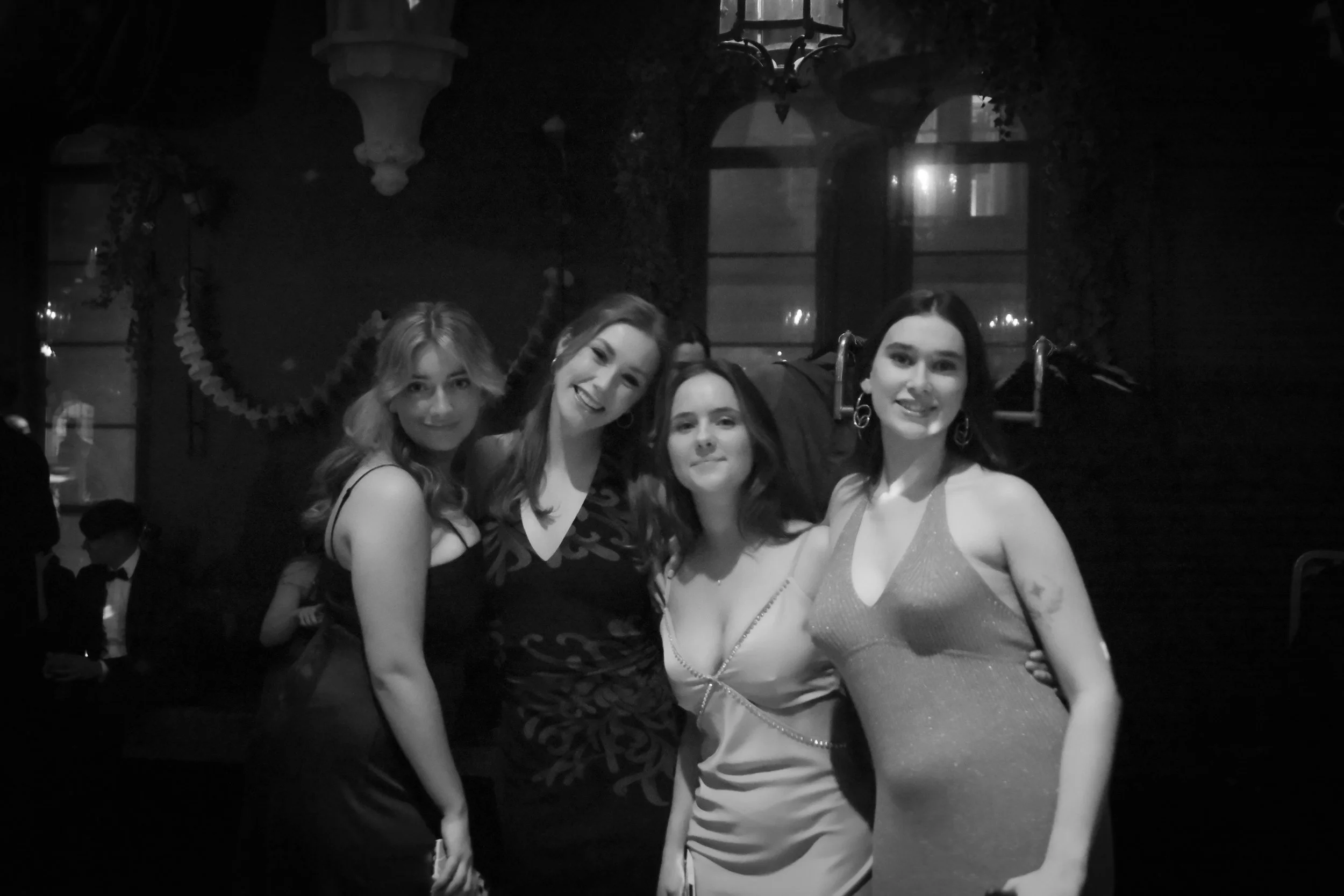 Four women dressed in formal attire, posing and smiling at a social event in a dimly lit indoor setting.