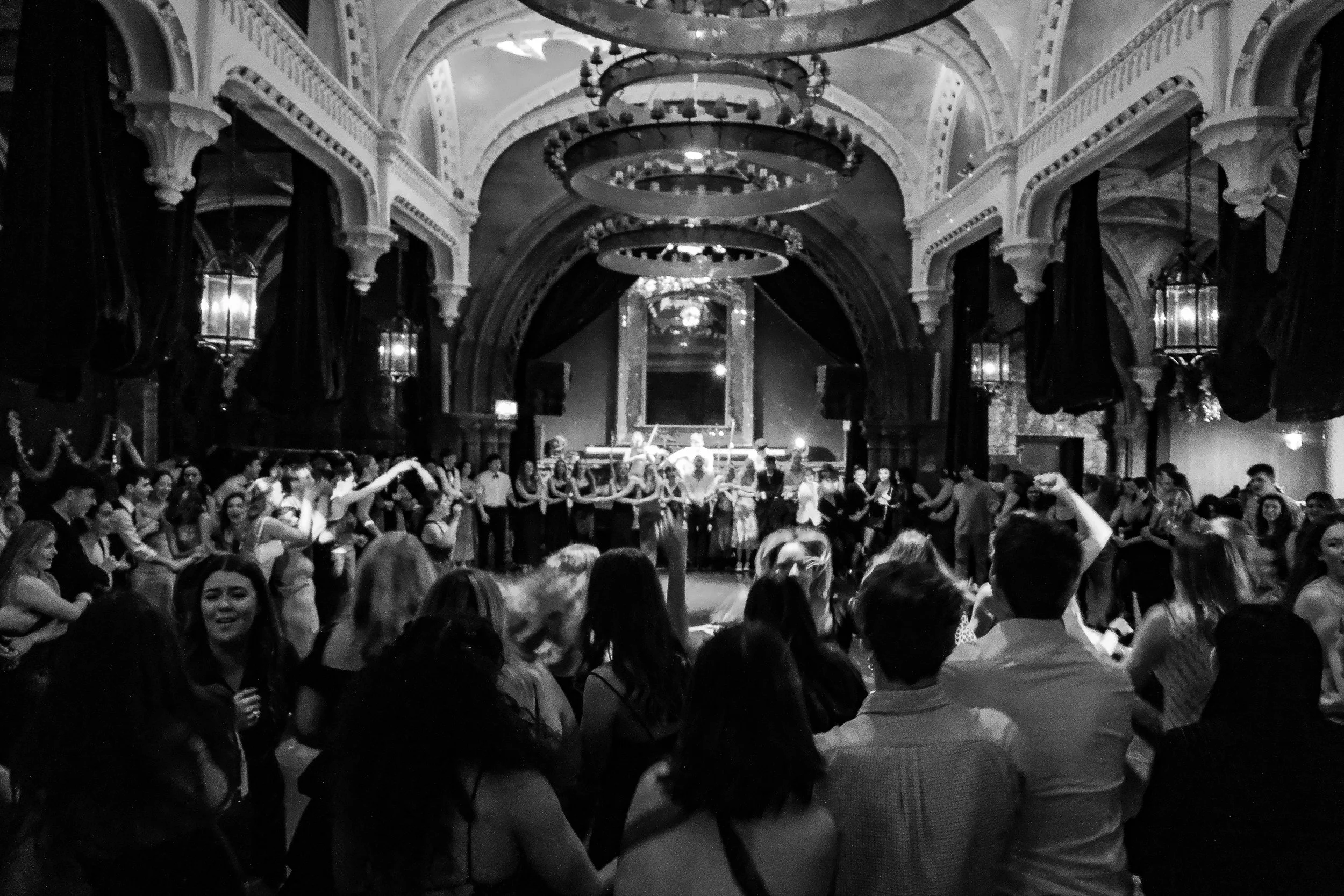 People dancing and socializing in a large ornate hall with high ceilings, chandeliers, and dark curtains during a festive event.