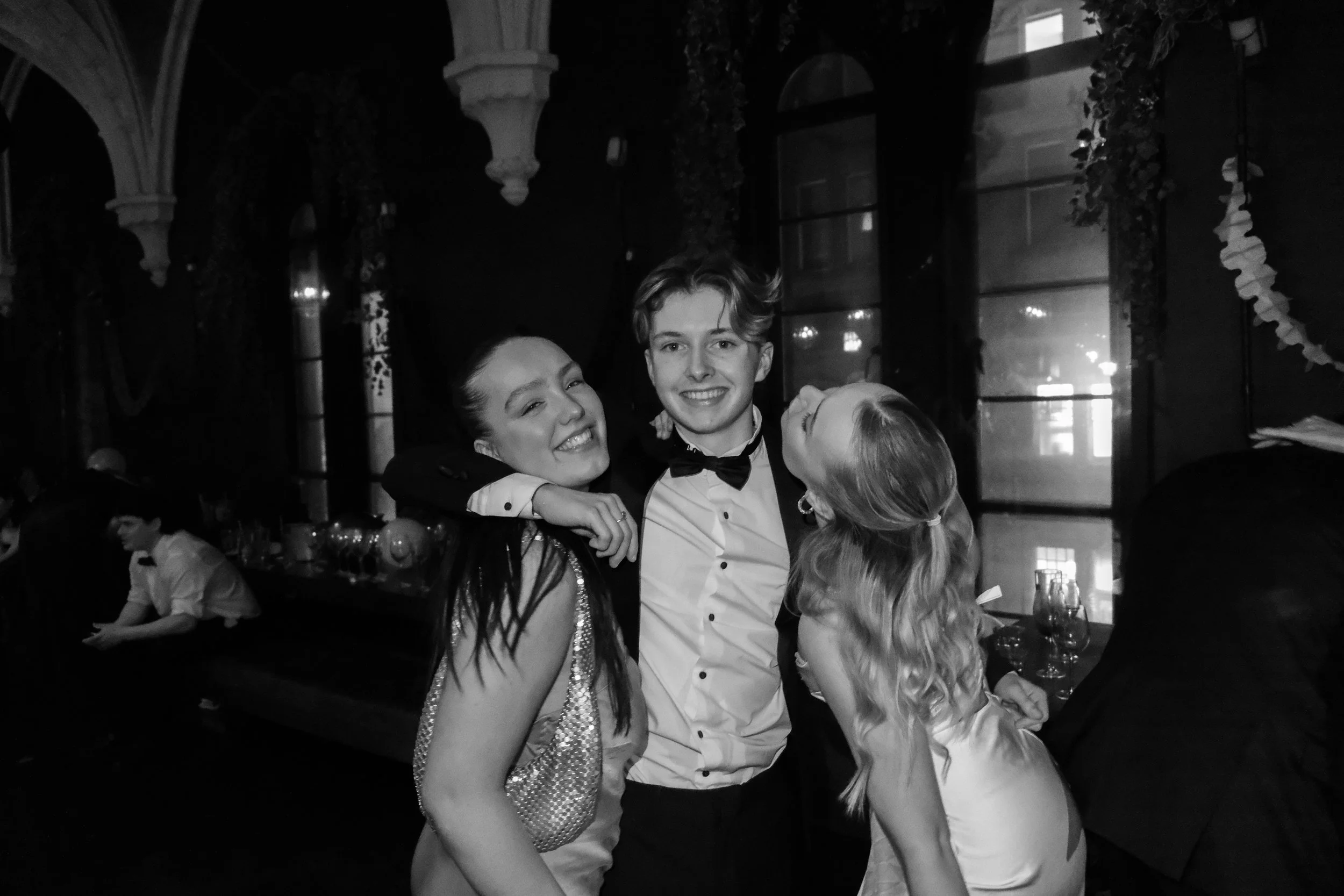 Black and white photo of three young adults at a formal event. They are smiling and laughing, dressed in elegant attire. The person in the middle is wearing a tuxedo with a bow tie, while the women on each side are in evening dresses. The background 
