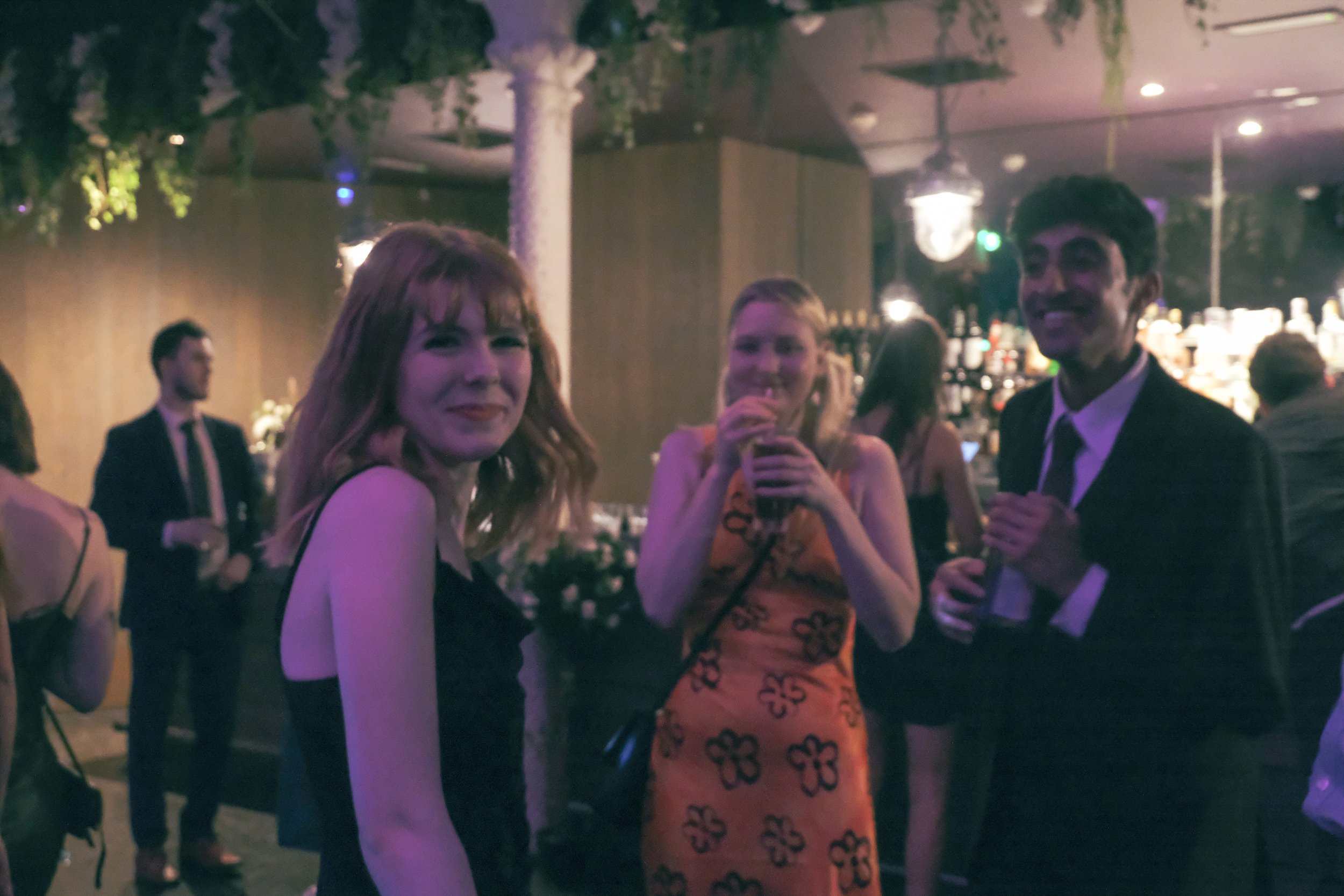 People at a social event, three are clearly visible in the foreground: a woman with red hair in a black dress, a woman in a sleeveless orange dress with black floral patterns holding a drink, and a man in a suit with a tie, smiling at the camera. Bac