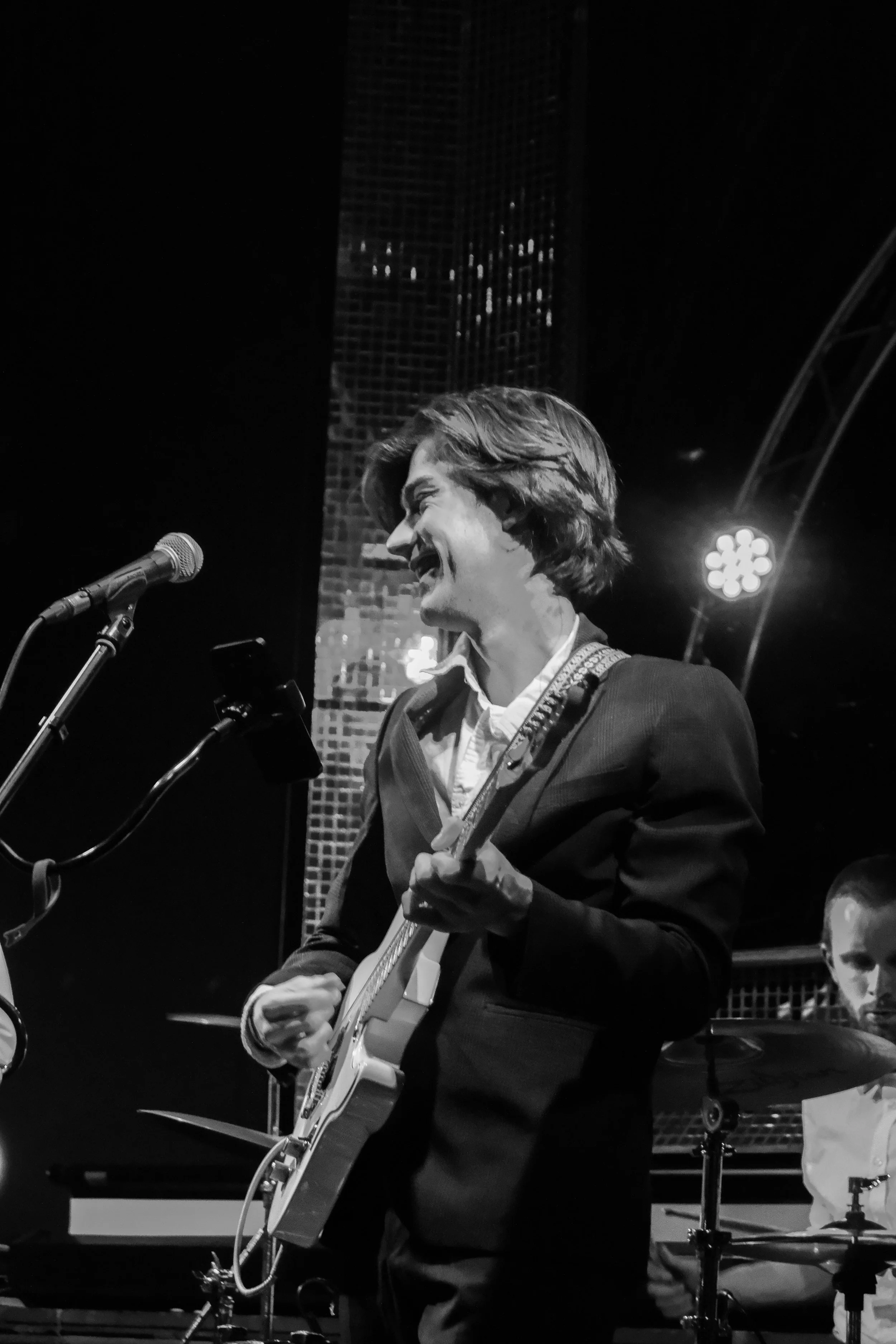 A man playing guitar on stage, smiling, with a drummer in the background, black and white photo.