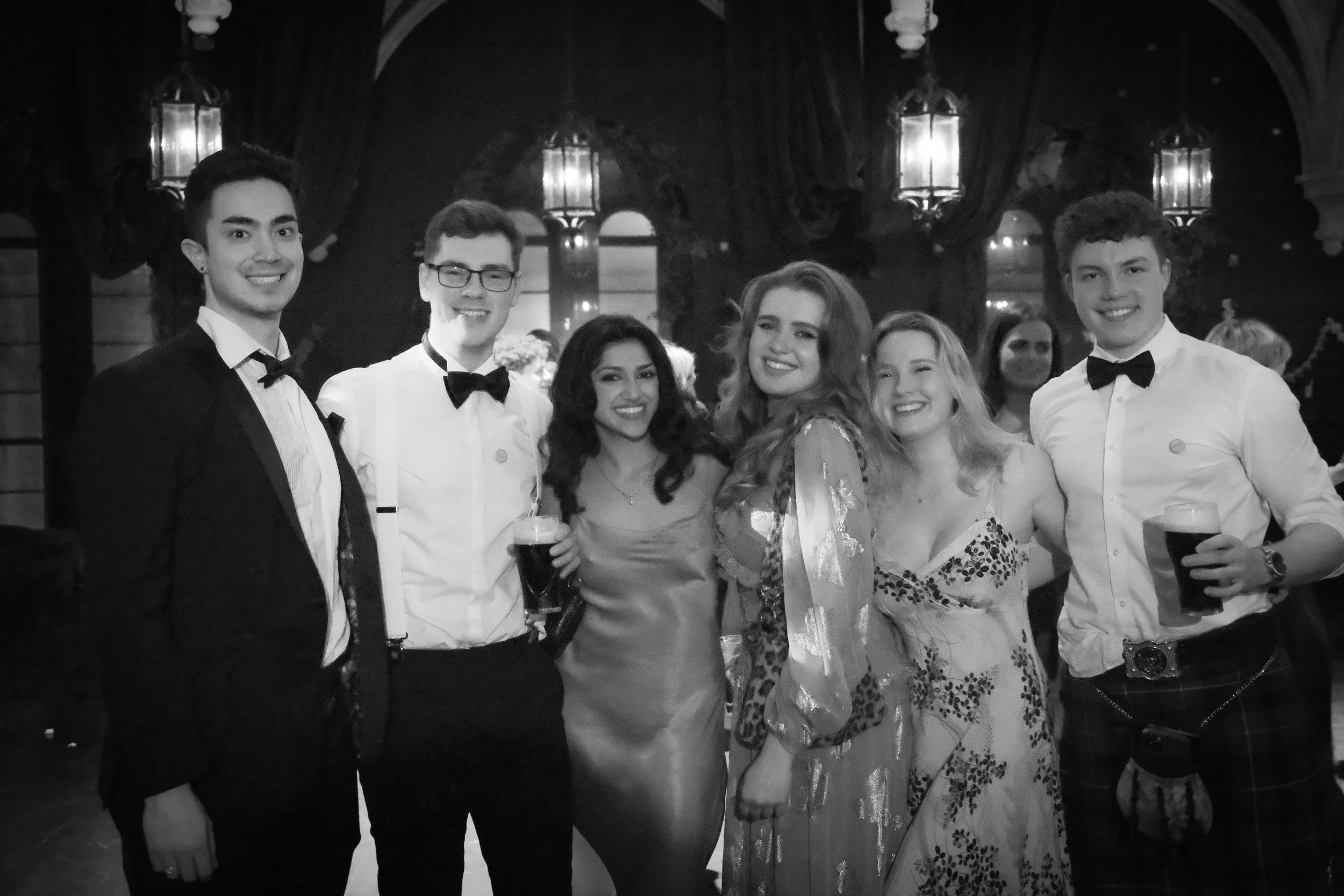 Group of six young adults in formal attire smiling together at a party or celebration, with ornate lighting and drapery in the background.