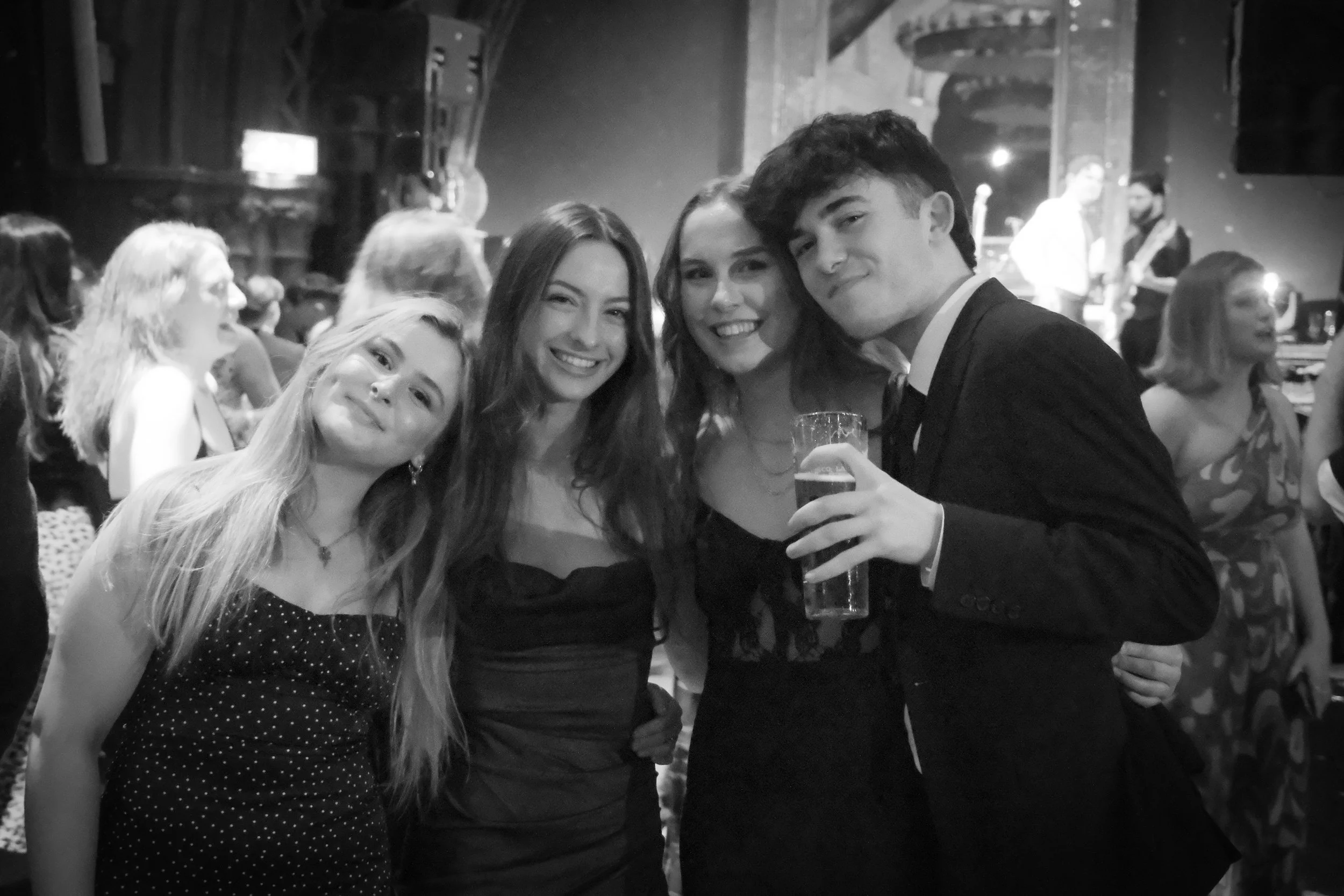 Group of four friends smiling at a party, with a band playing on stage in the background. One person is holding a glass of beer.