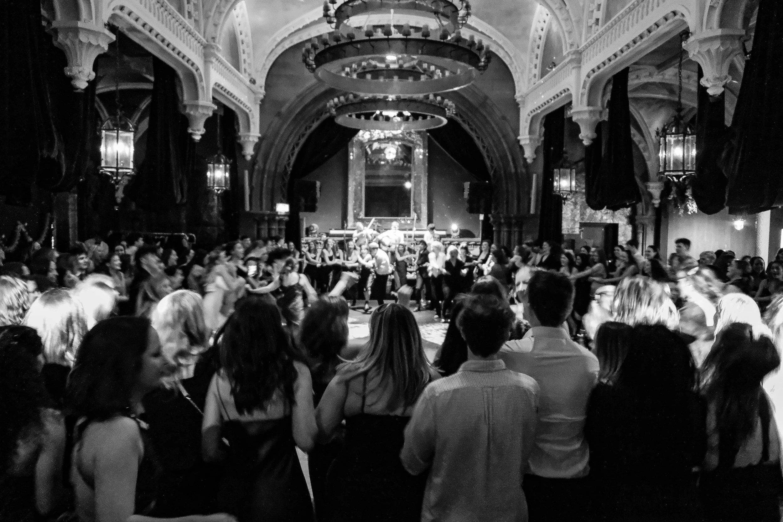 A black and white photo of a lively party or dance event inside a grand, ornate hall with high ceilings, intricate architectural details, and chandeliers. Many people are dancing in the center, while others are watching from the perimeter.