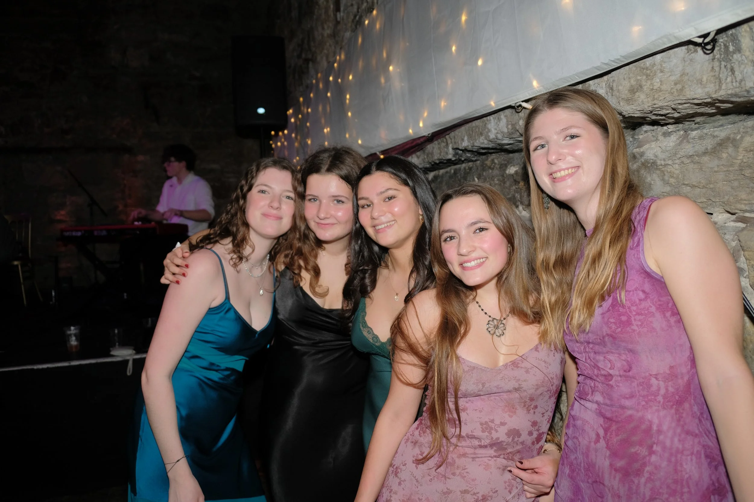 Group of five young women in elegant dresses smiling and posing together indoors, with a stone wall and string lights in the background.