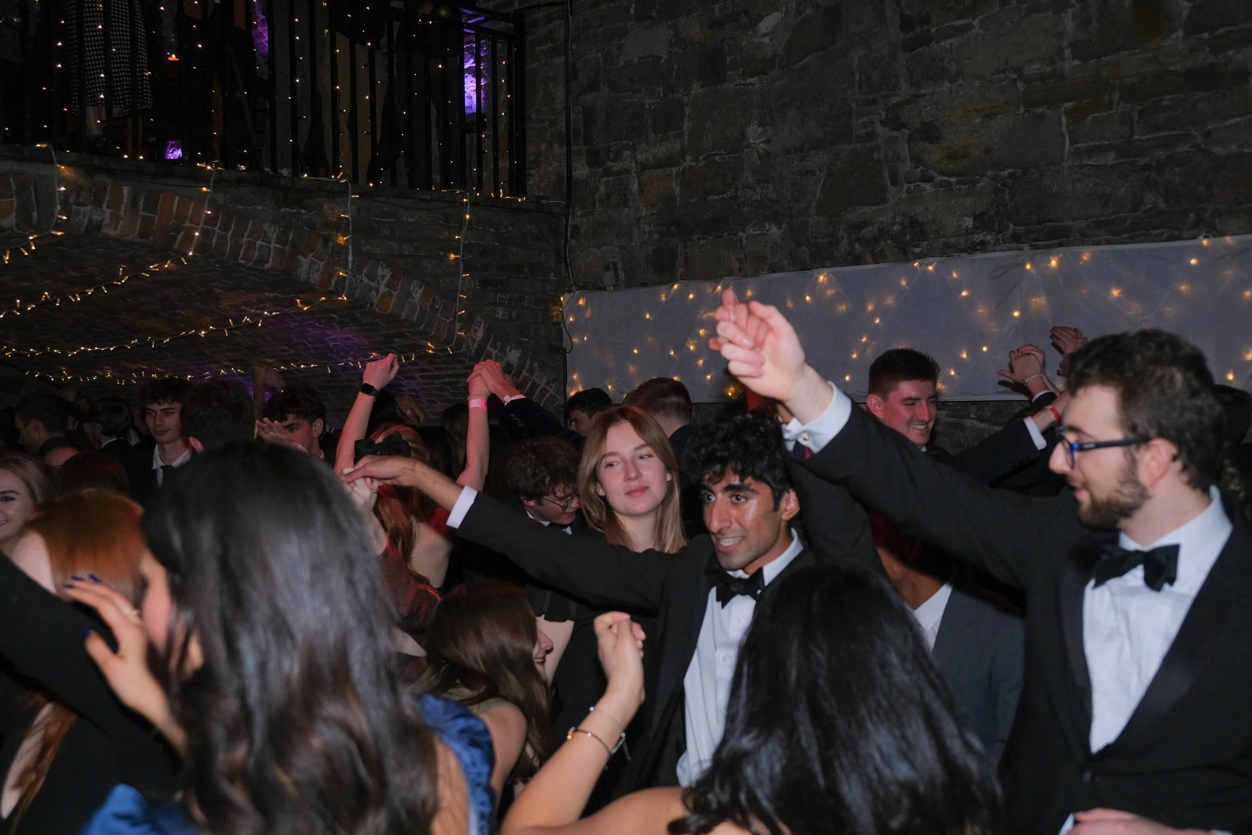 Young people dancing and celebrating at a party, dressed in formal attire with tuxedos, in a dimly lit venue decorated with string lights and a stone wall.