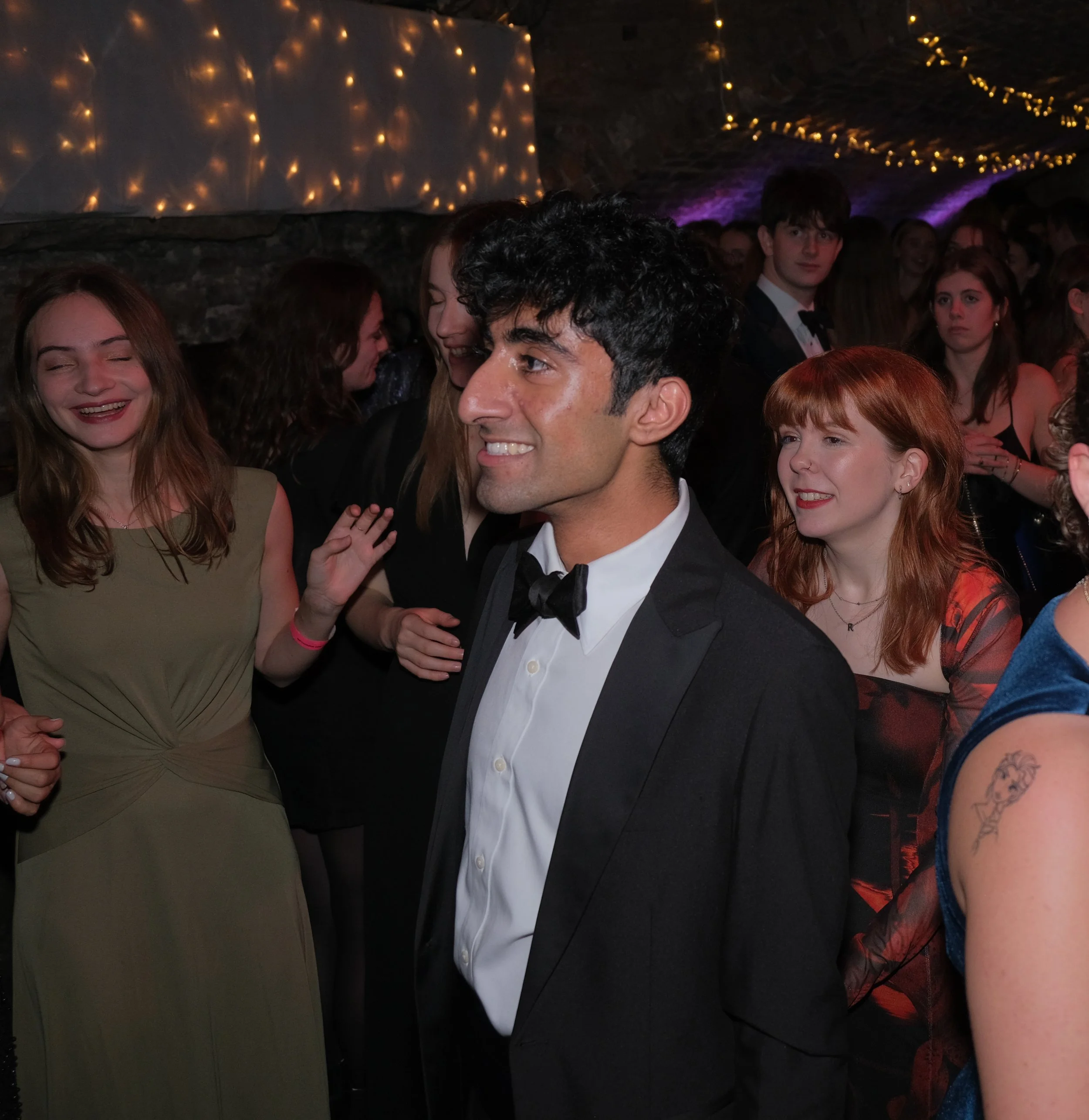Man in tuxedo smiling at a party or dance event with women around him.