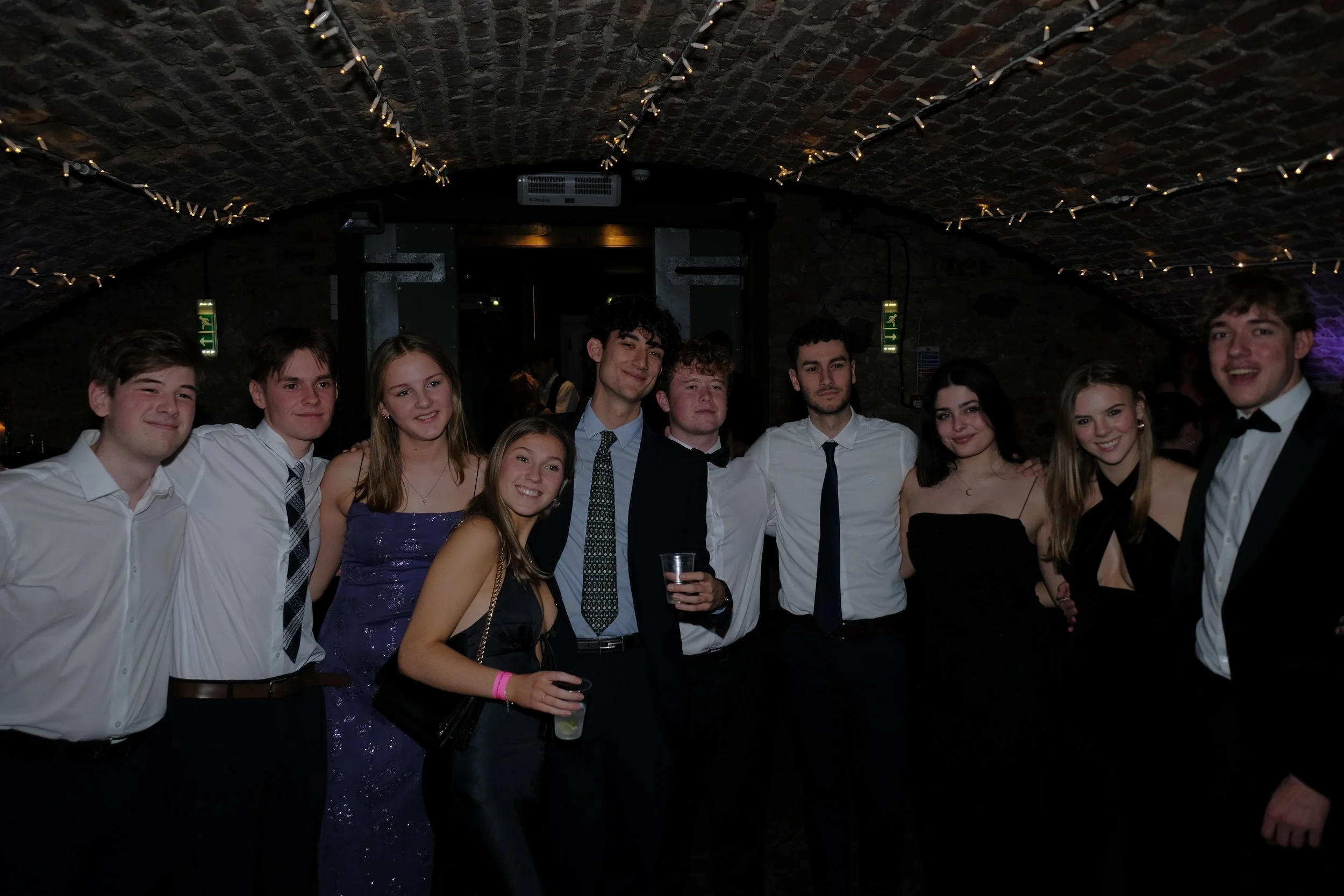 Group of ten young people dressed in formal attire at a party or event in a dimly lit venue with string lights overhead.