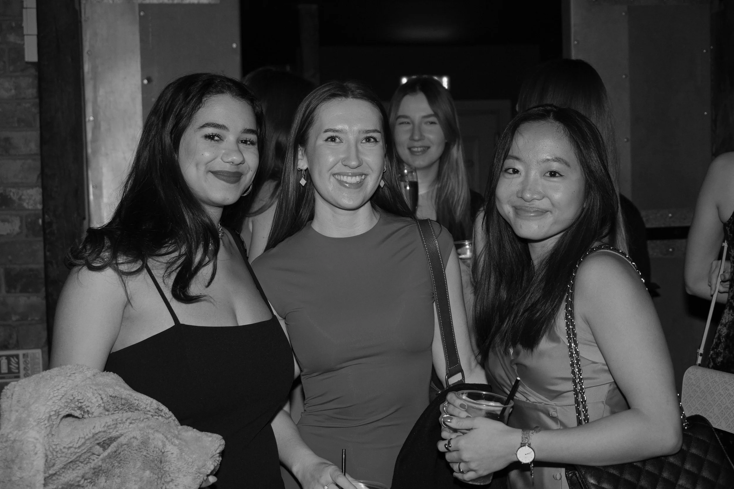 Four women smiling at a social event, standing closely together in a dimly lit indoor venue.
