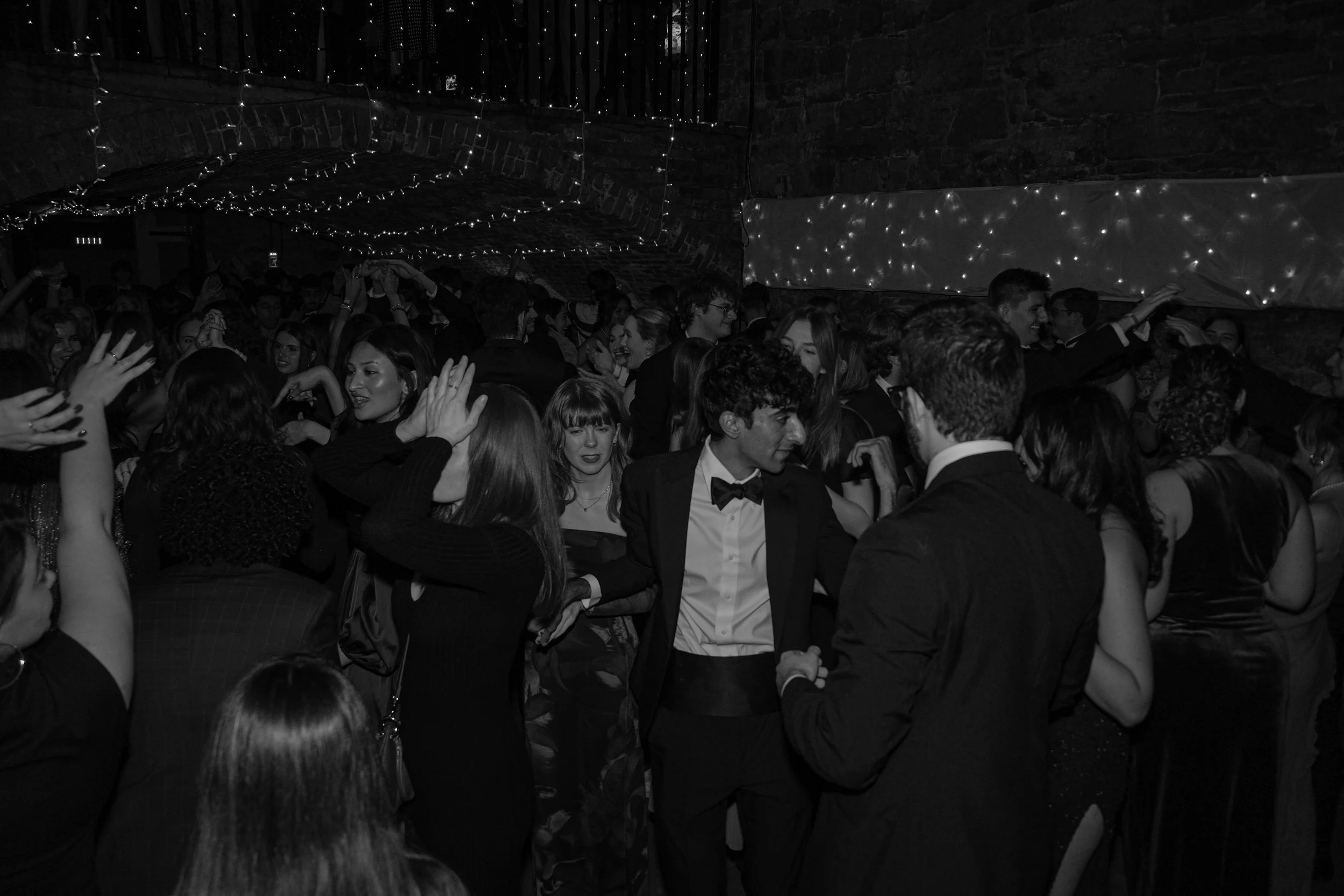 People dancing and socializing at a party with string lights and a brick wall background.