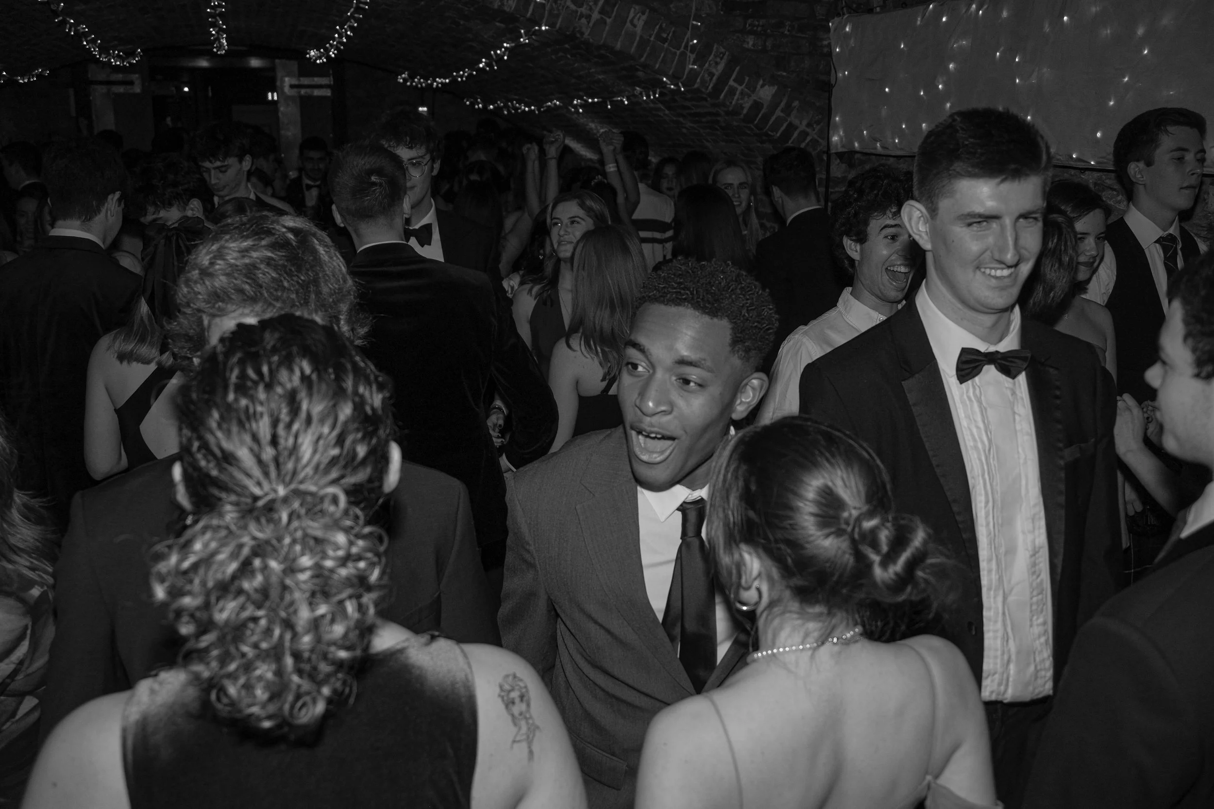 People at a formal dance or party, wearing tuxedos and dresses, in a decorated venue with string lights.
