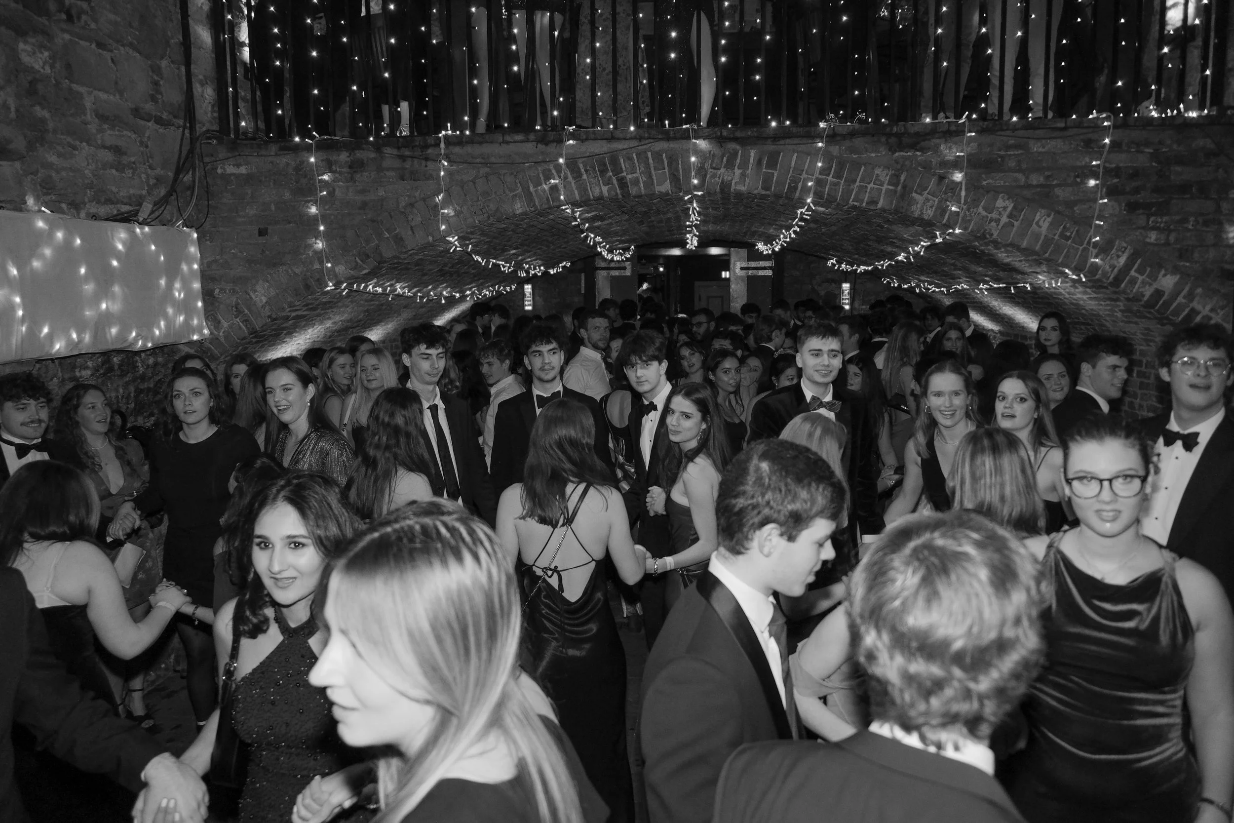 A black and white photo of young men and women dancing at a formal party in a brick venue decorated with string lights.