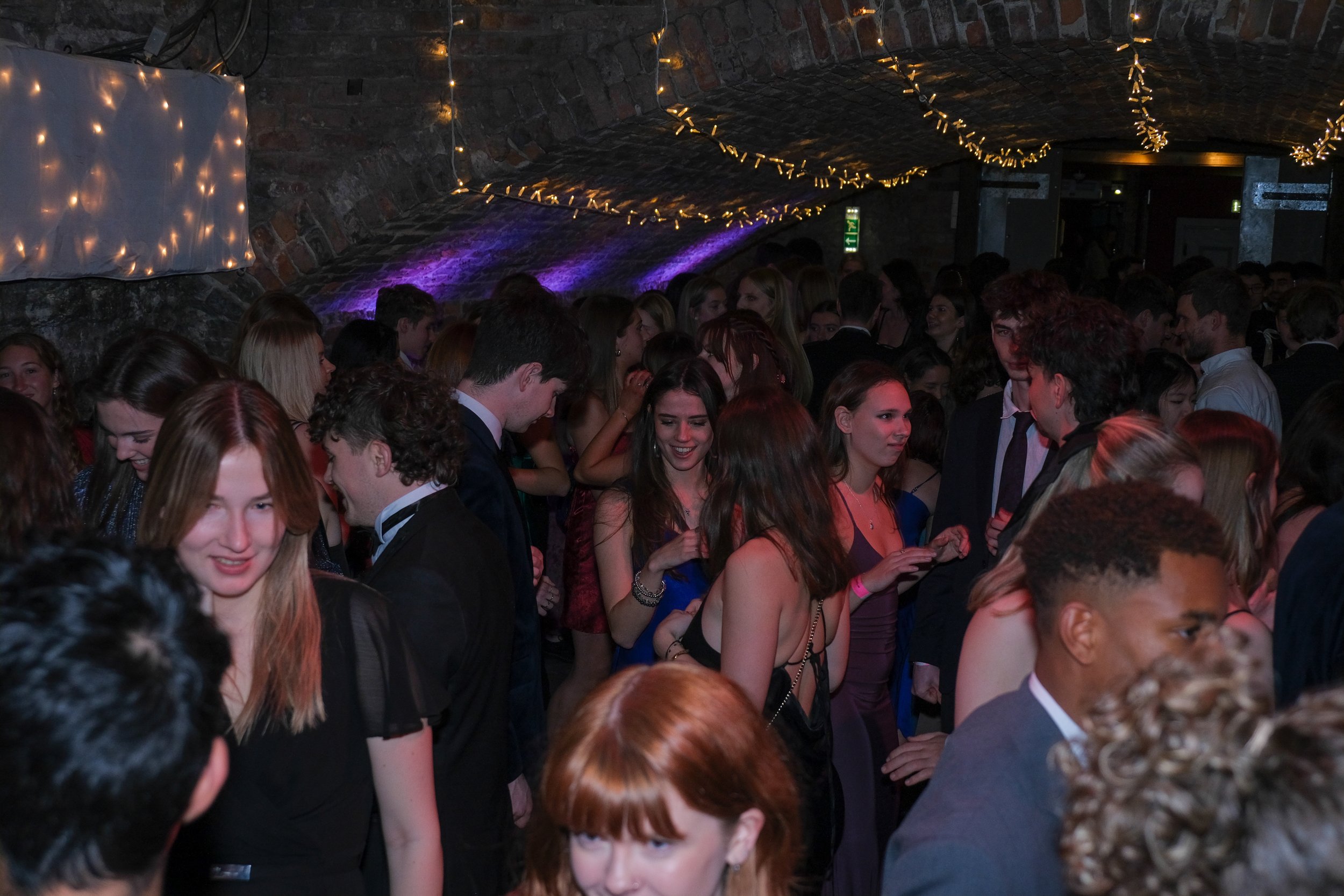 Crowd of young adults at a party or dance event under a brick arch ceiling with string lights and purple lighting.