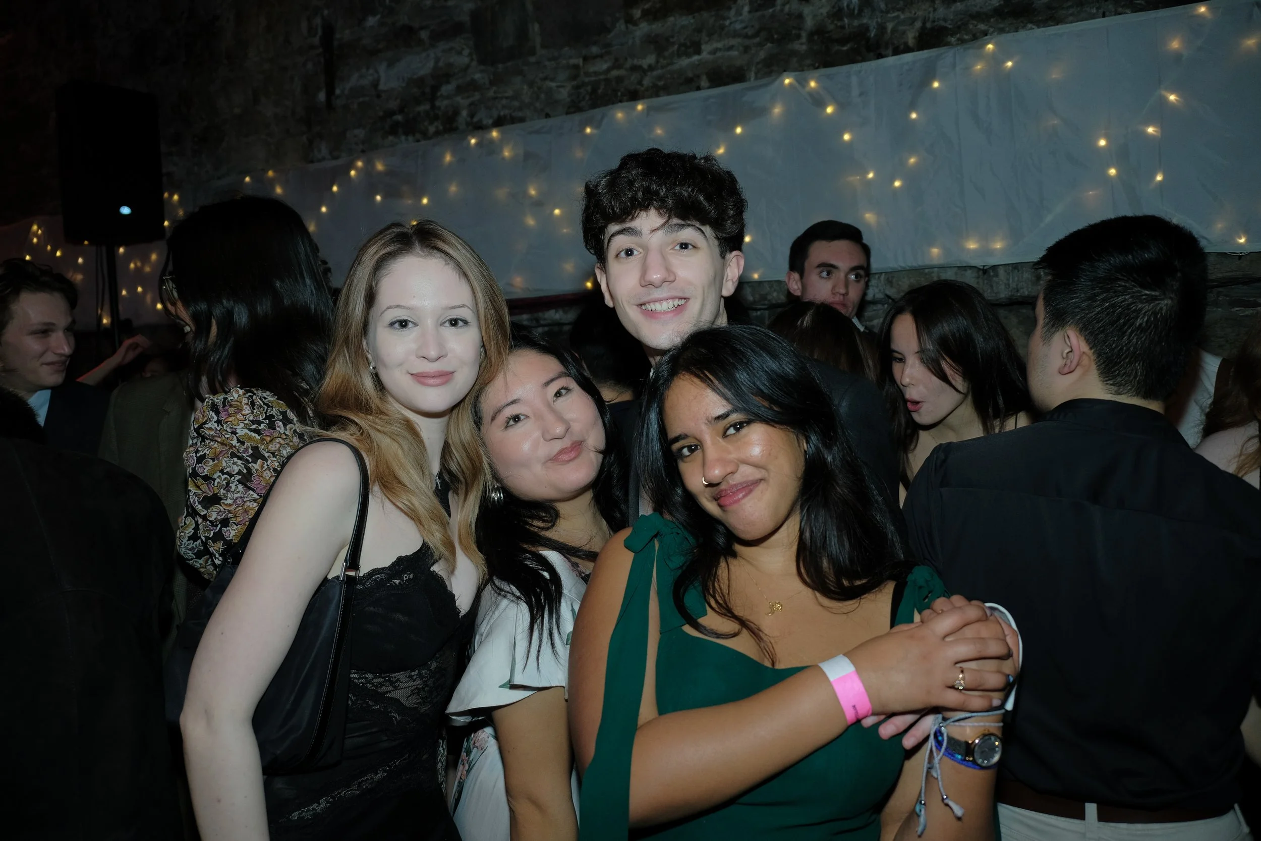 Group of six young adults at a party, smiling and posing for the photo. The background has fairy lights and a bar or wall with stones.