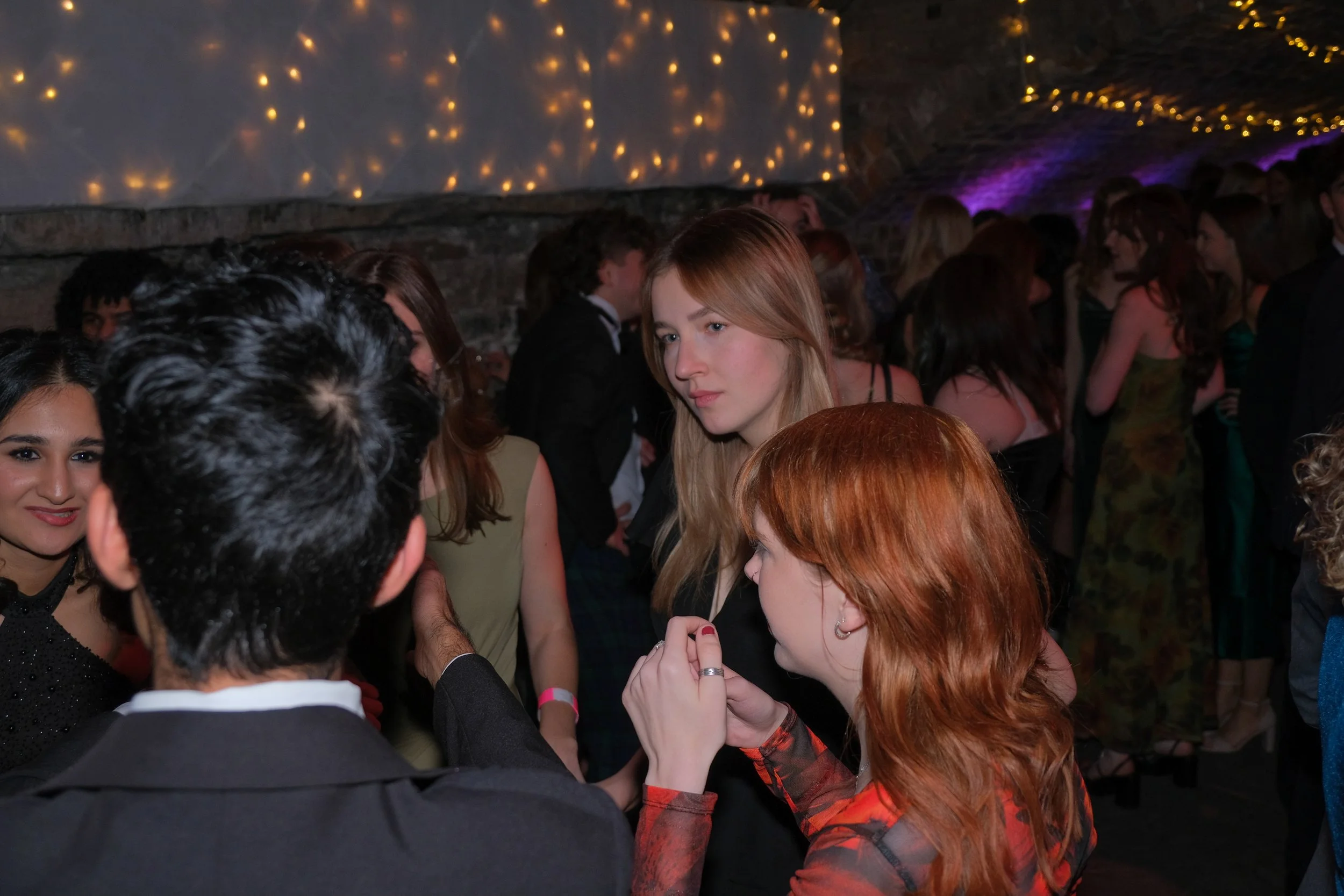 People dancing at a party or event in a dimly lit room with string lights overhead.