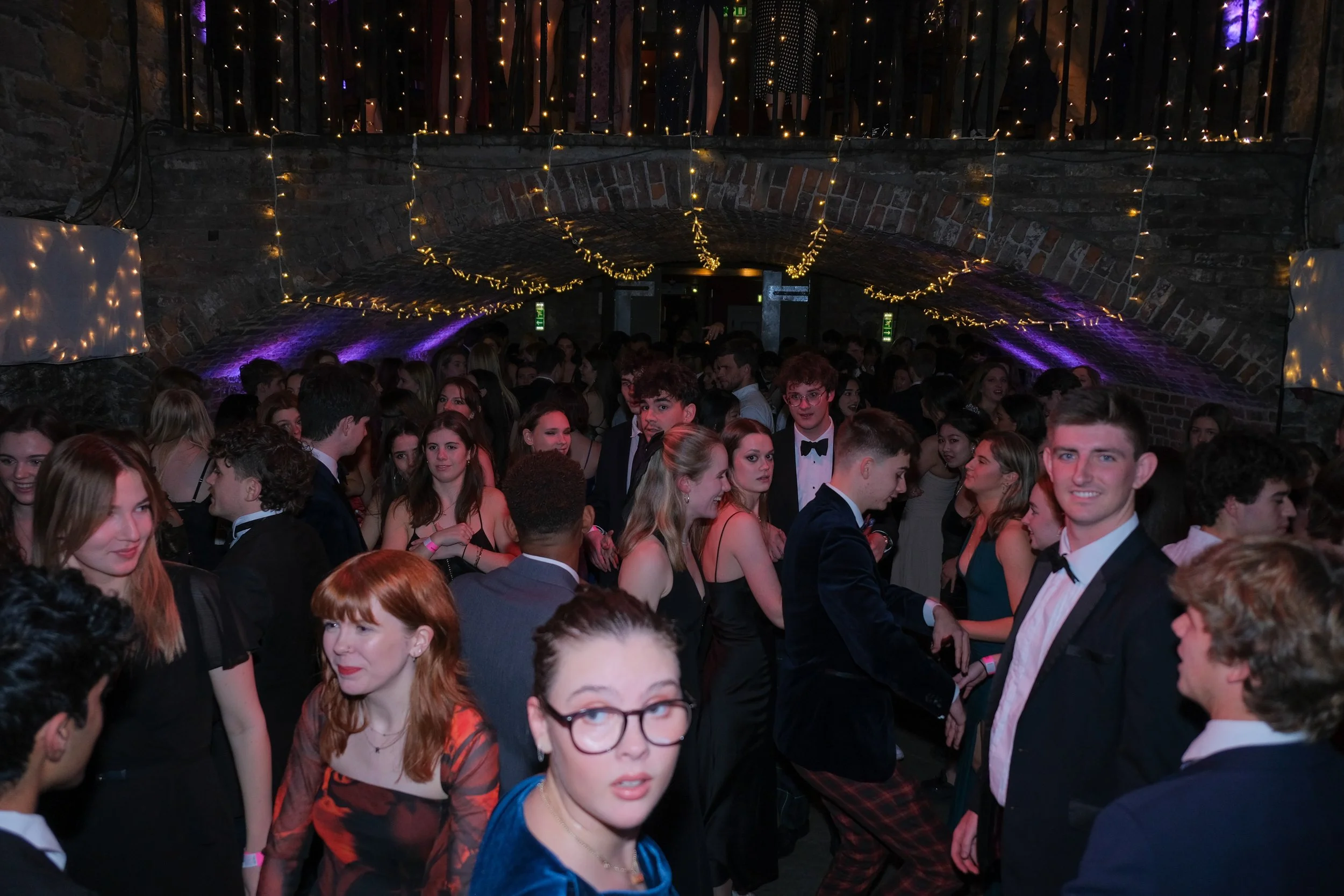 Crowd of young people dancing at formal event under brick arch with string lights.