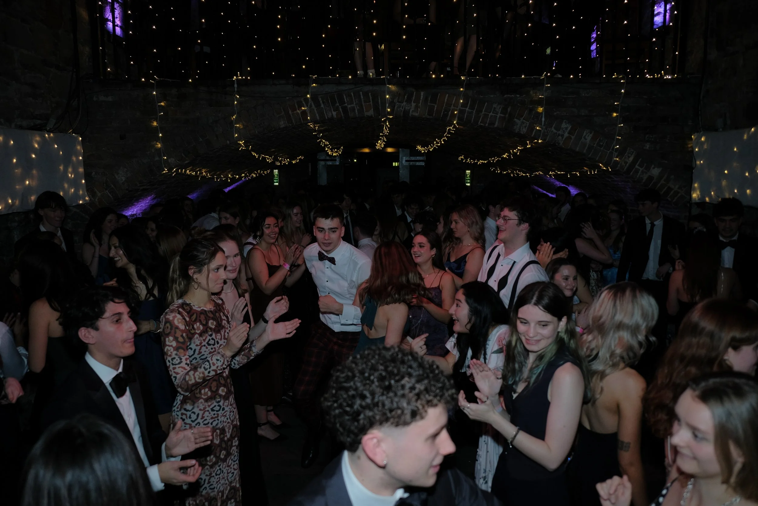 Group of young people dancing and socializing at a party with string lights and a brick arched ceiling.
