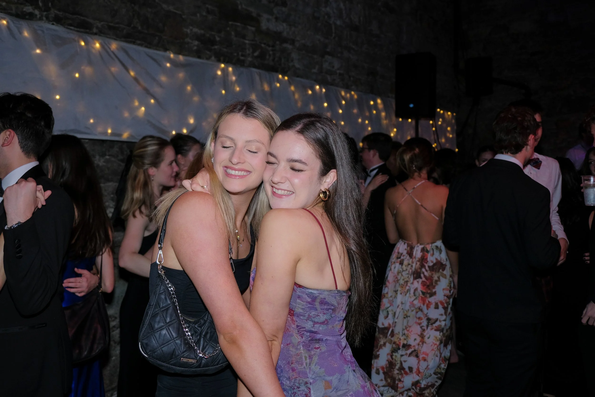 Two women hugging and smiling at a party or dance event, with other people dancing and socializing in the background under string lights.
