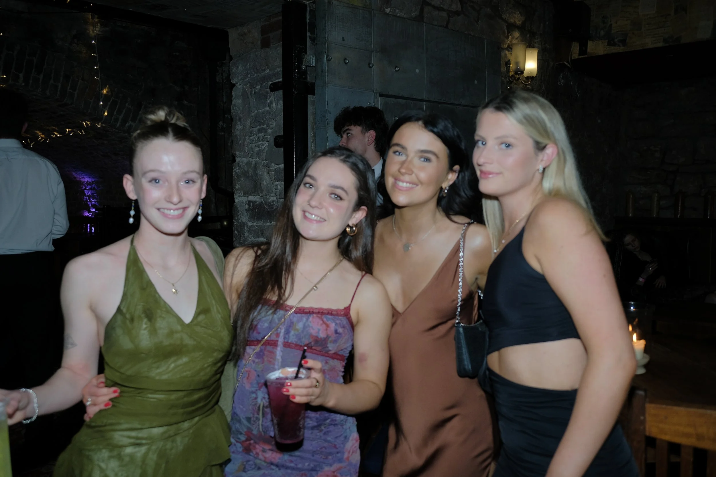 Four women posing together at a party or social gathering in a dimly lit venue with stone walls.
