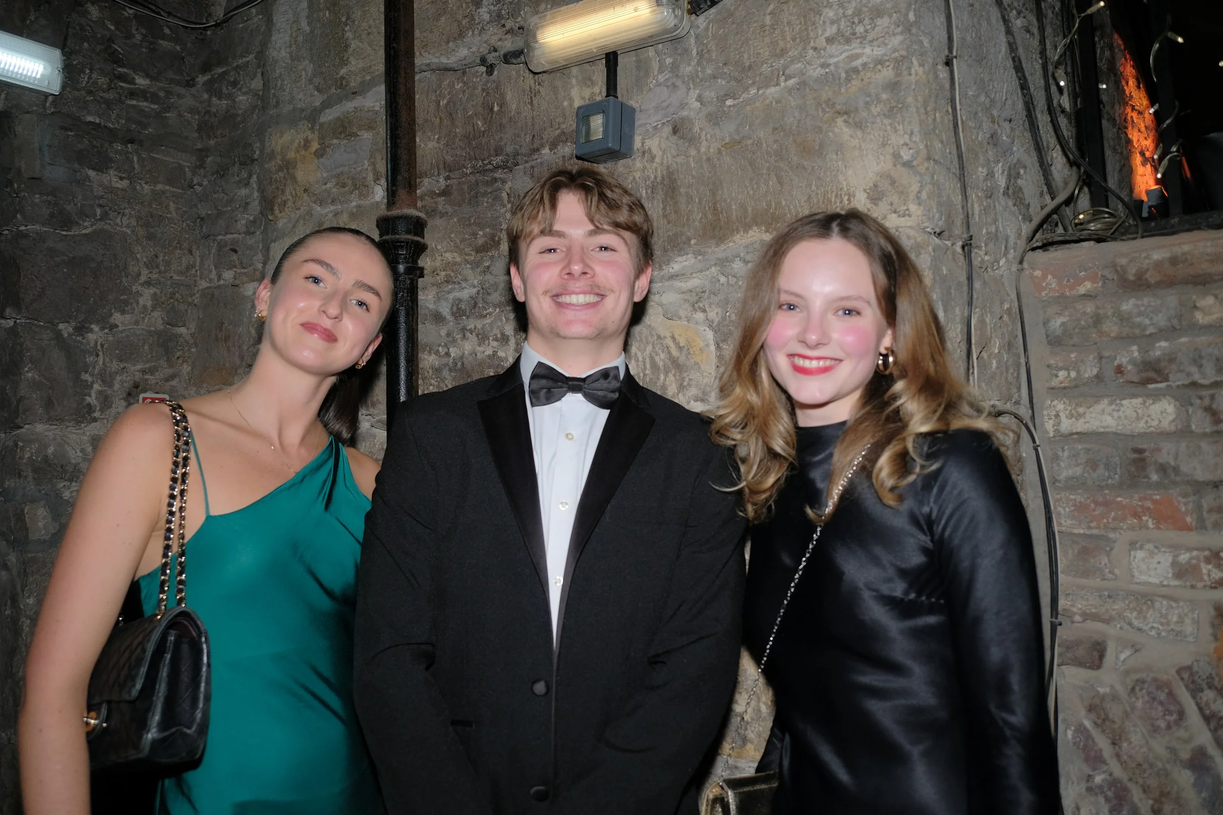 Three young adults dressed in formal attire, two women and one man, smiling at the camera against a stone wall background.