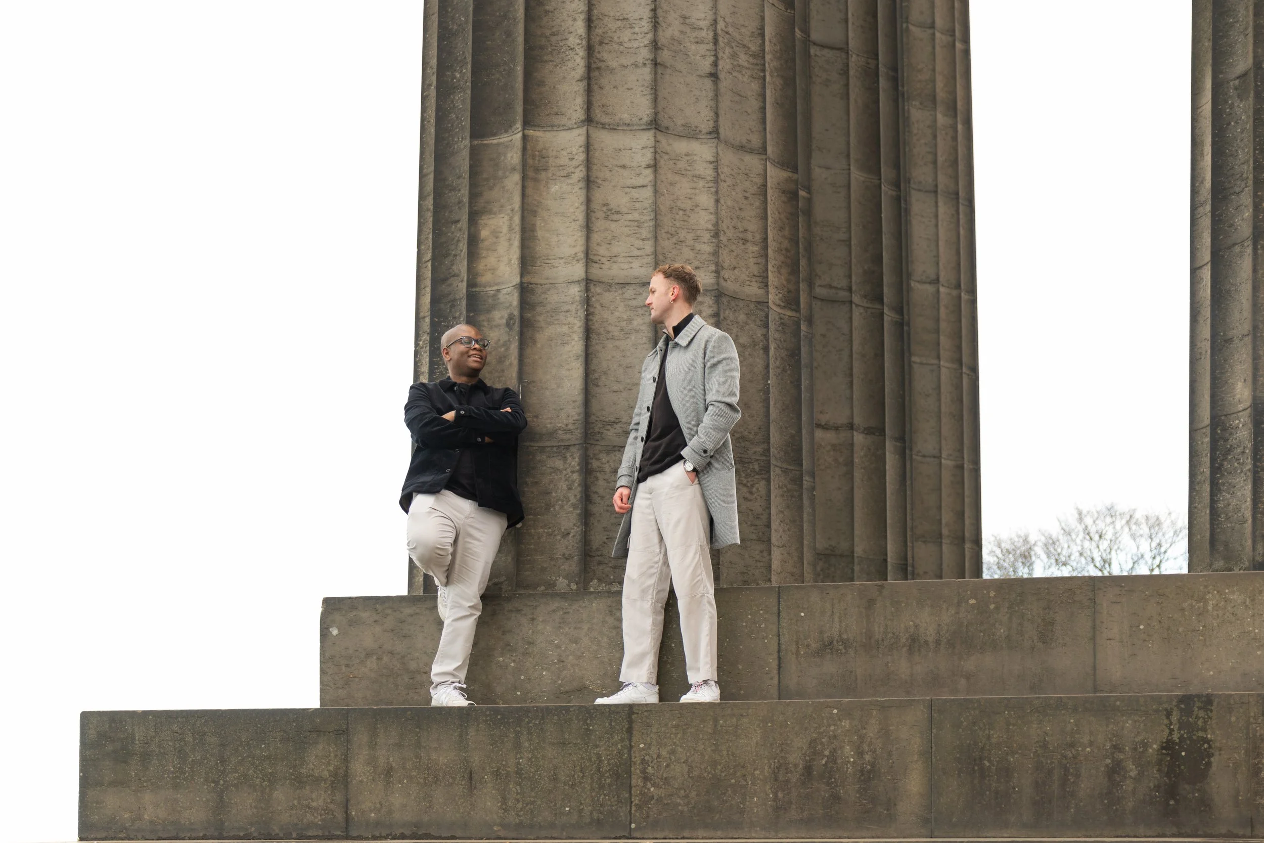 Two people talking and smiling in front of a large, tall stone column with a cloudy sky in the background.