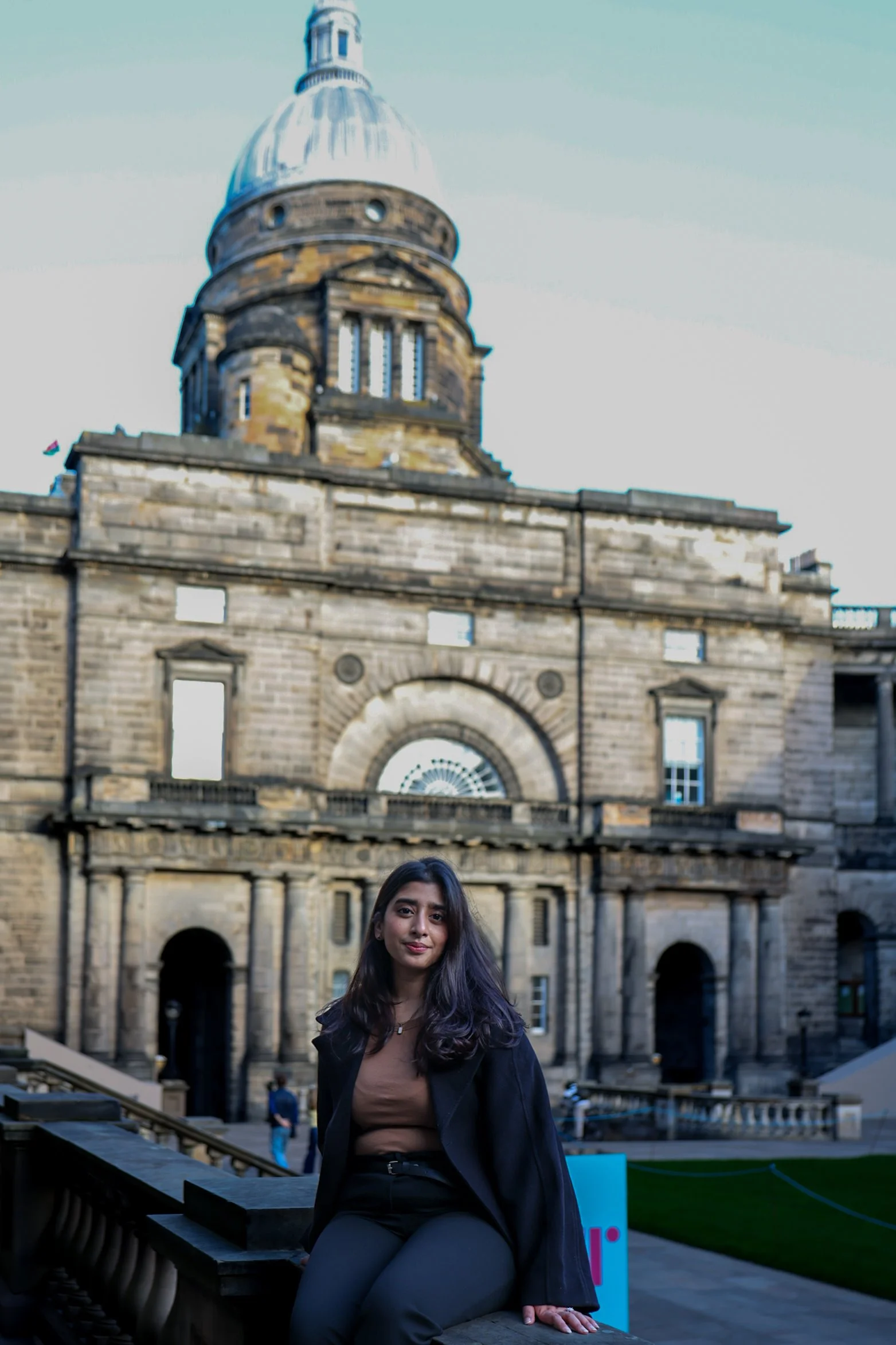 A woman sitting on a stone ledge in front of an old building with a large dome. The building appears to be a historical or government structure made of stone, with a mix of rounded and rectangular windows and architectural details.