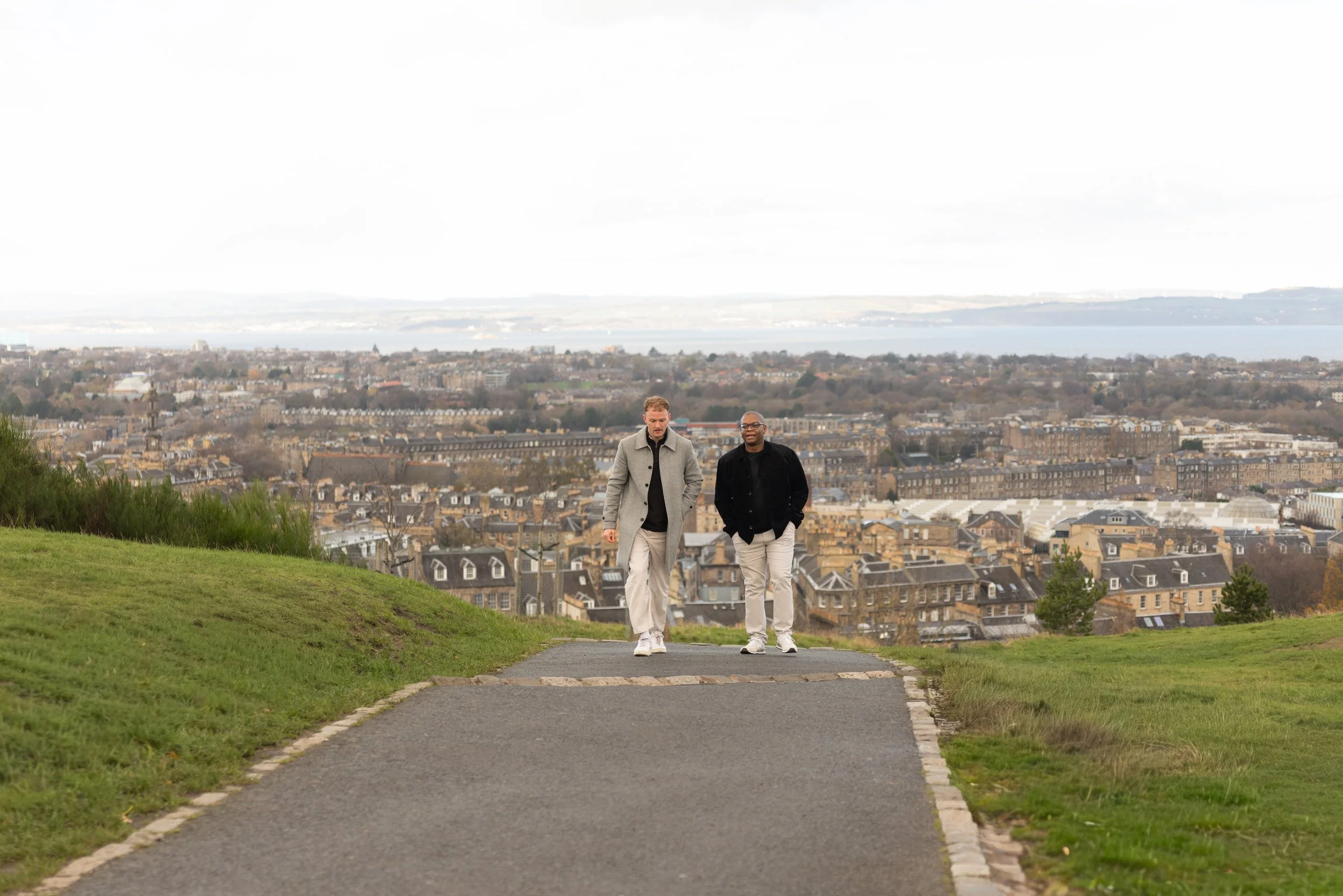 Two men walking on a paved path towards a cityscape with a body of water in the background, surrounded by grassy areas.